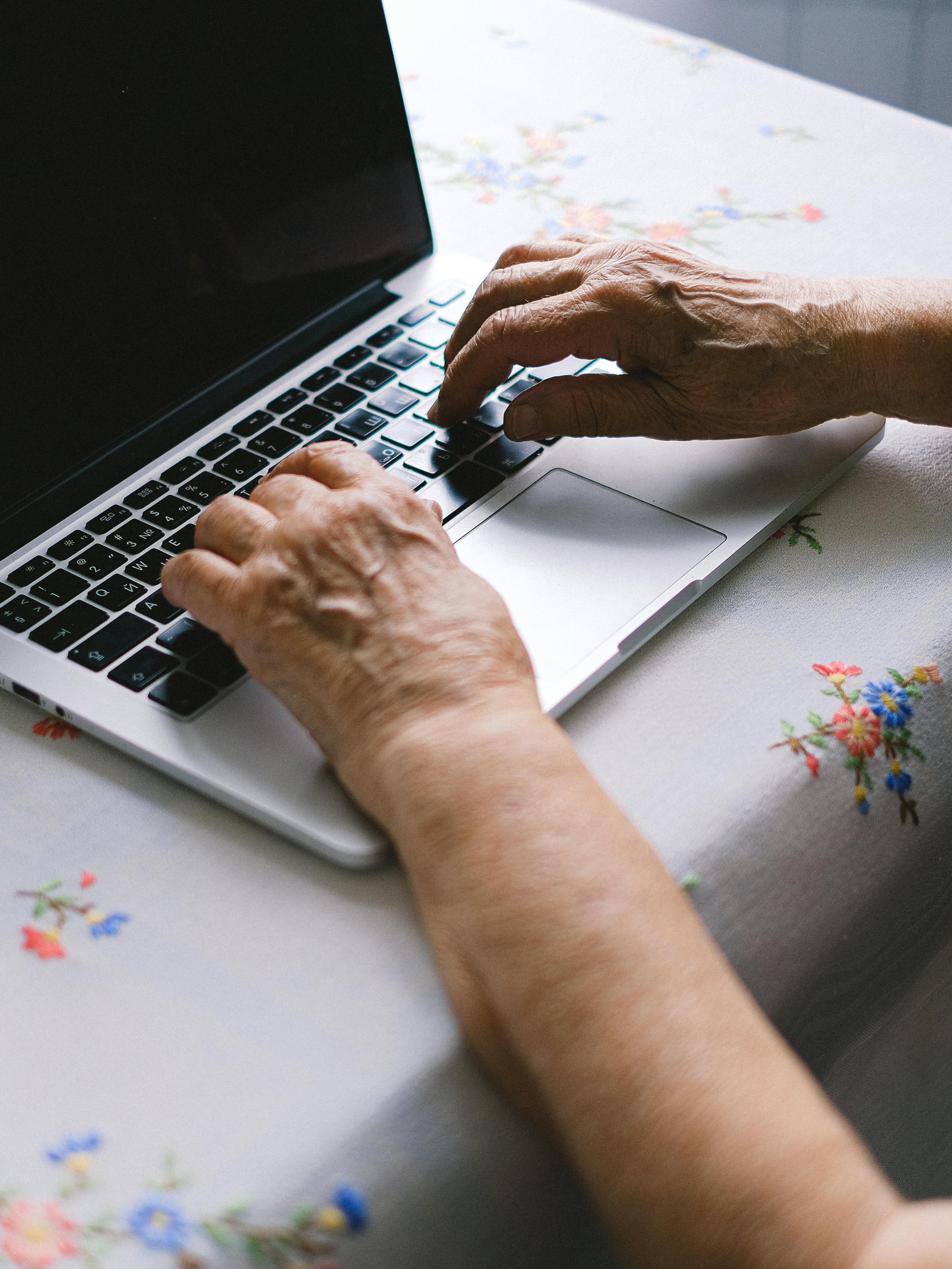 Hände einer älteren Person tippen auf einer Laptop-Tastatur auf einem blumigen Tischdecken.