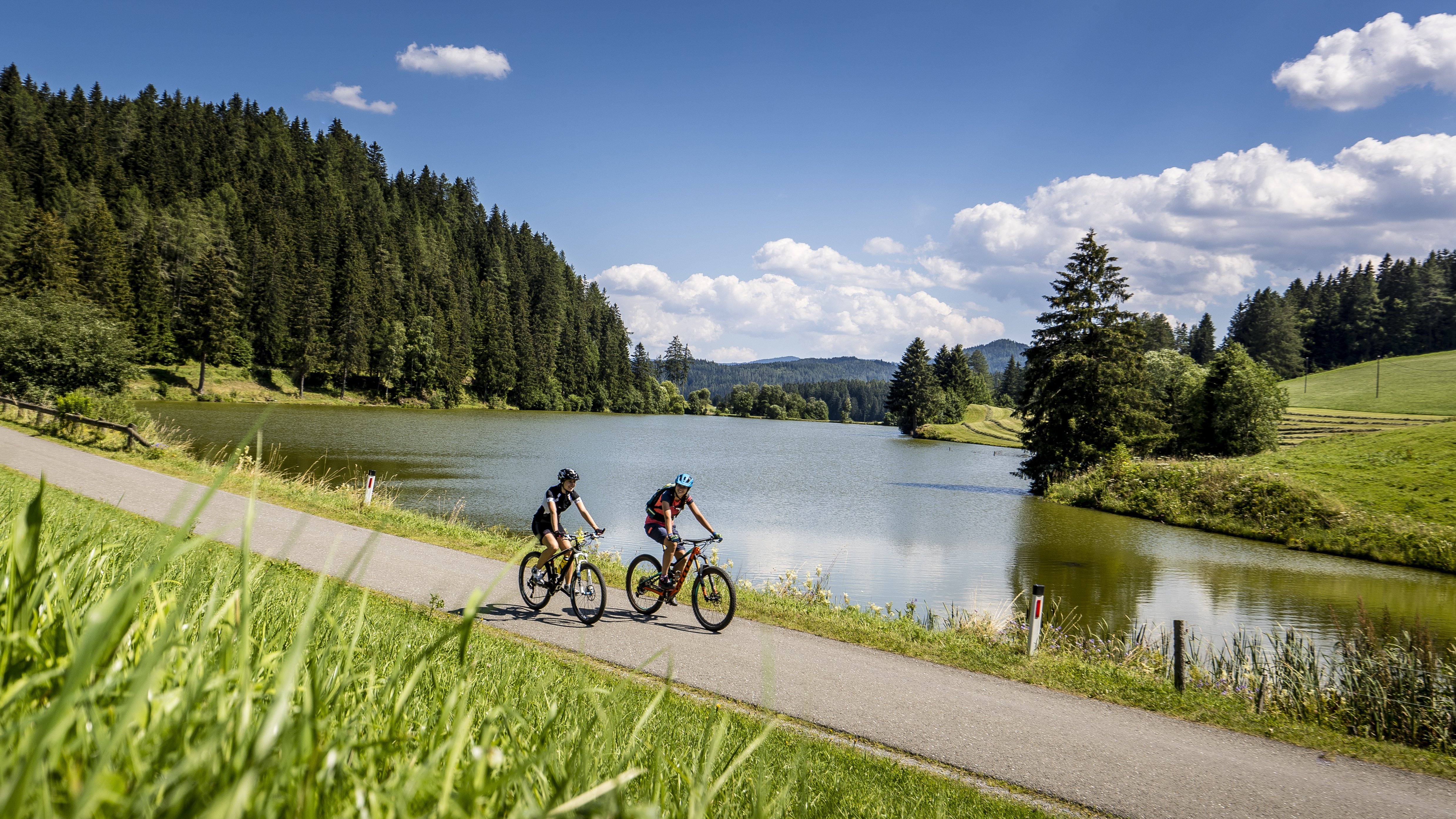 Mountainbike Abenteuer in der Natur Zwei Radfahrer auf einem Weg neben einem See, umgeben von Bäumen und Grünflächen.