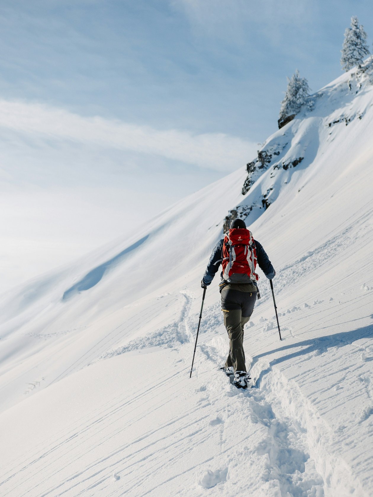 Eine Person wandert mit Schneeschuhen auf einem verschneiten Berghang unter blauem Himmel.