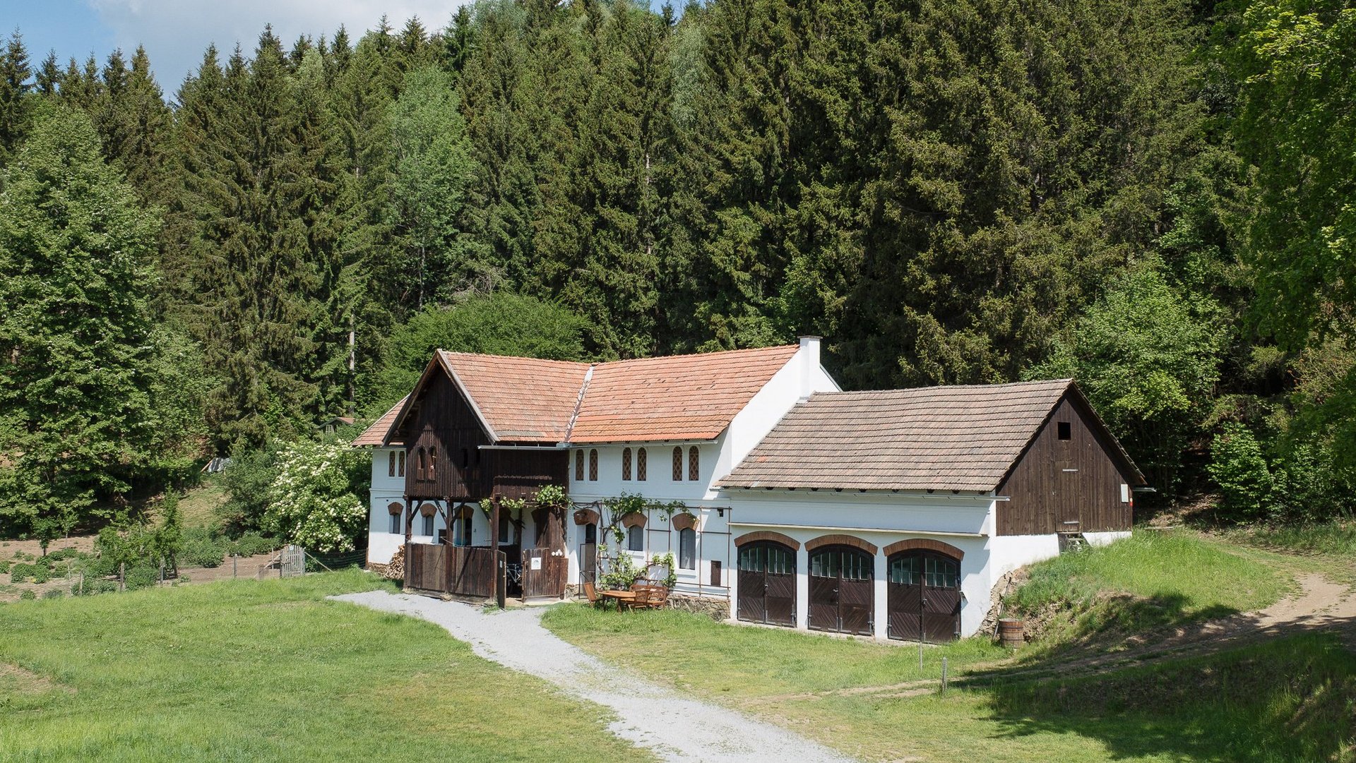 Ein Landhaus umgeben von Bäumen und üppigem Grün vor einem Waldhintergrund. Es vermittelt ein Gefühl von Ruhe und Natur.