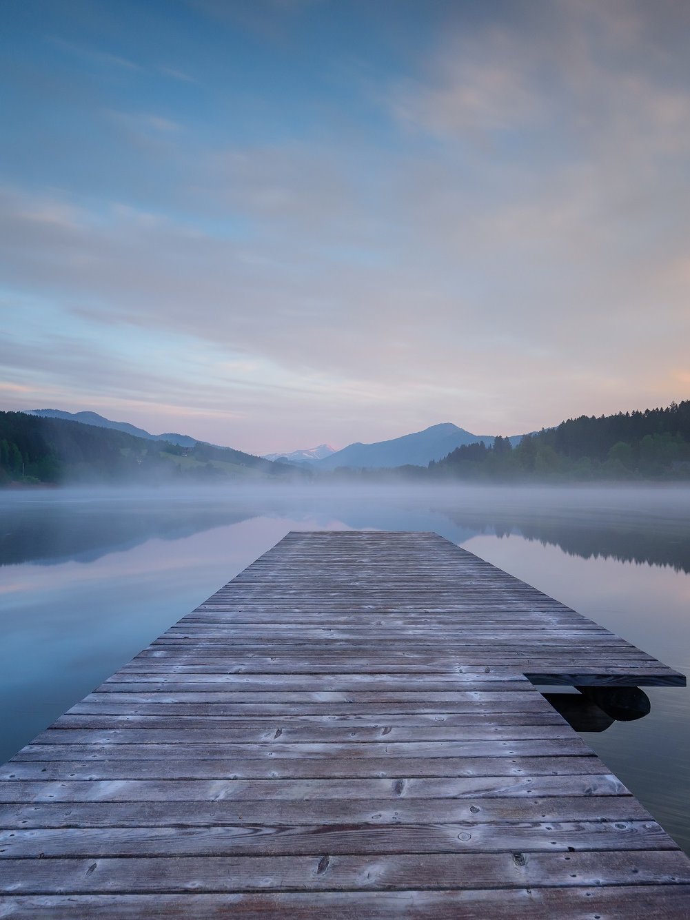 Morgendliche Stille am Furtner Teich Holzsteg führt in ruhigen See umgeben von nebligen Bergen bei Sonnenaufgang und sanfter Morgenstimmung