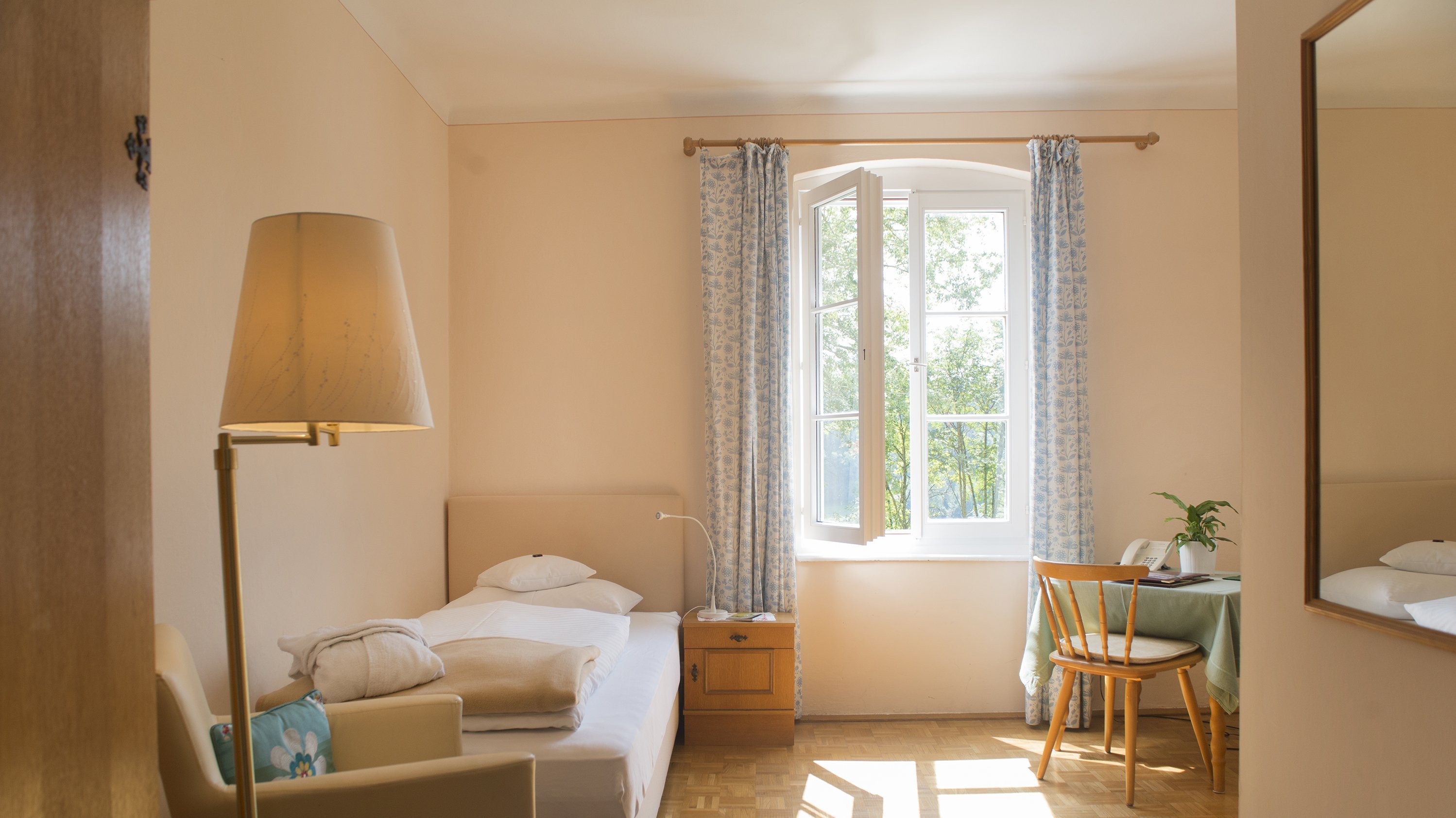 Gemütliches Einzelzimmer mit natürlichem Licht Einzelzimmer mit einem Bett, Stuhl und Tisch am Fenster, beleuchtet von Tageslicht.