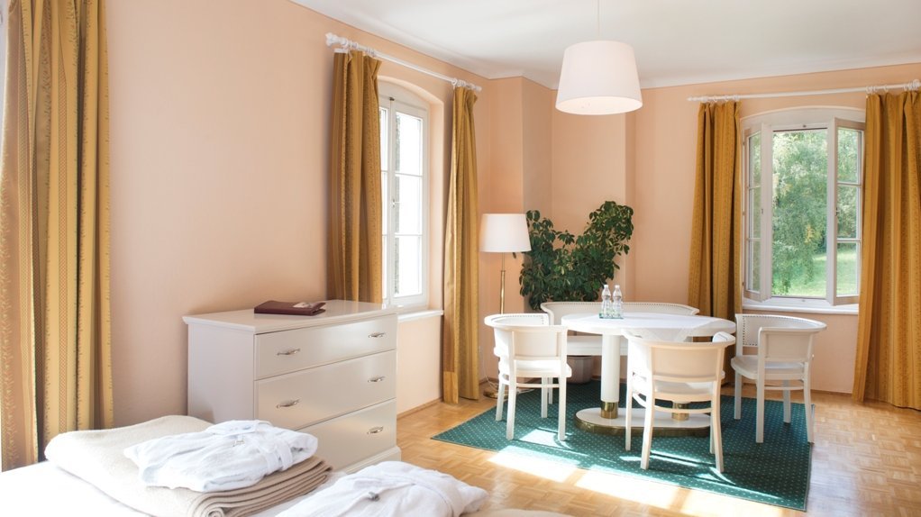 A well-lit bedroom featuring peach-colored walls, a dresser, and a small dining area by large windows.