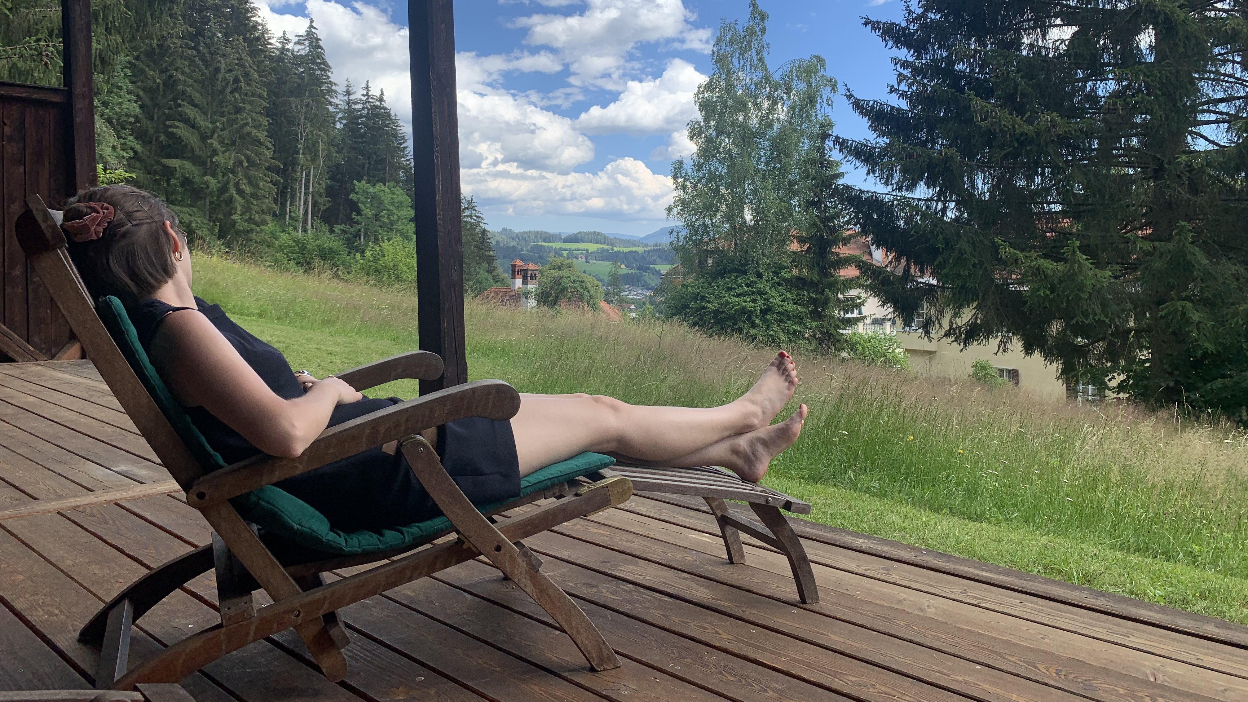 Person entspannt auf einem Holzliegestuhl mit Blick auf eine grüne Landschaft und bewölkten Himmel