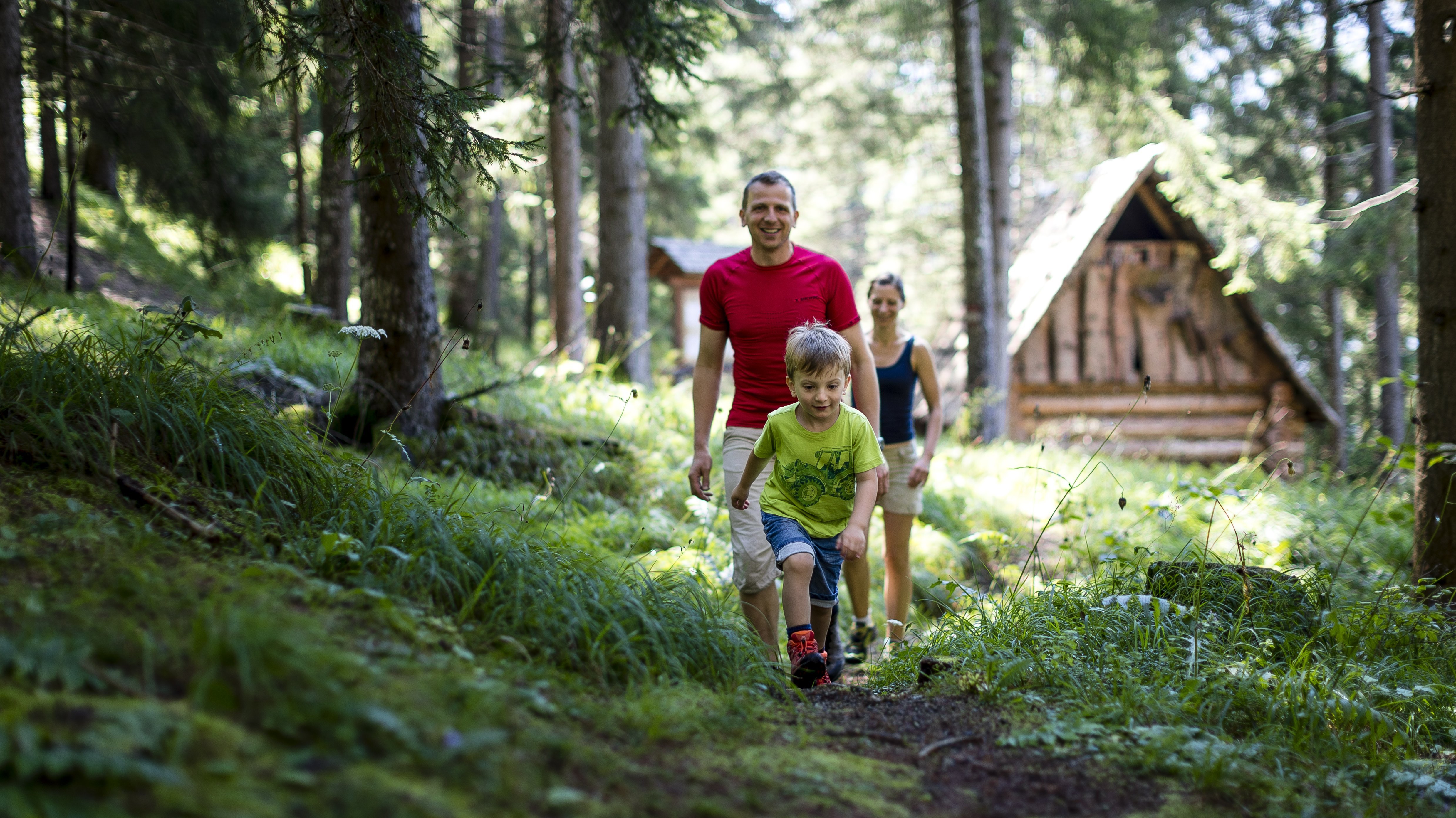 Familienwanderung im Wald Familie wandert durch einen bewaldeten Bereich mit einer Hütte im Hintergrund, ein Kind führt den Weg.