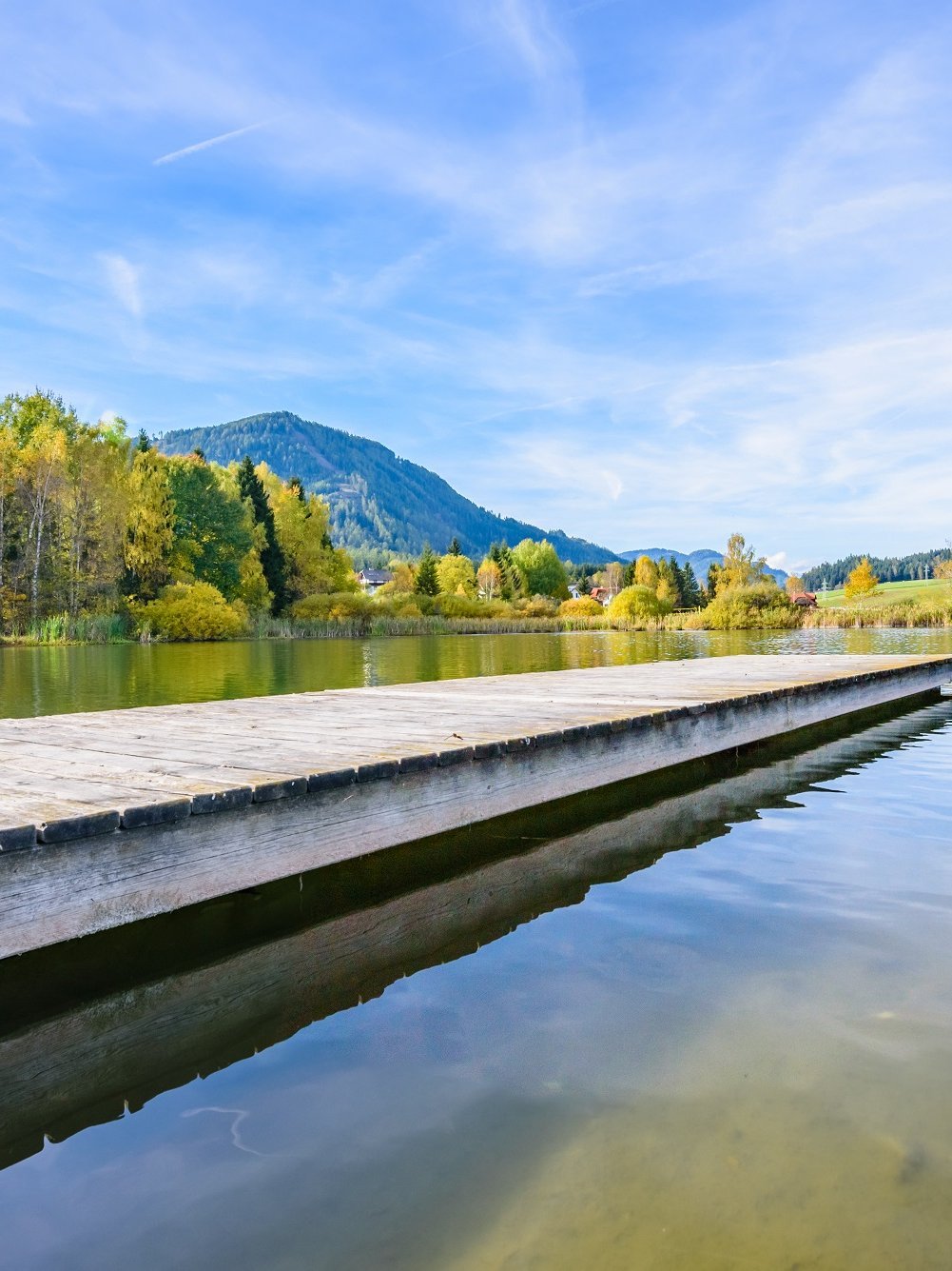Herbstliche Idylle am See Ruhiger See umgeben von Bäumen mit Herbstlaub und einem Holzsteg unter blauem Himmel.