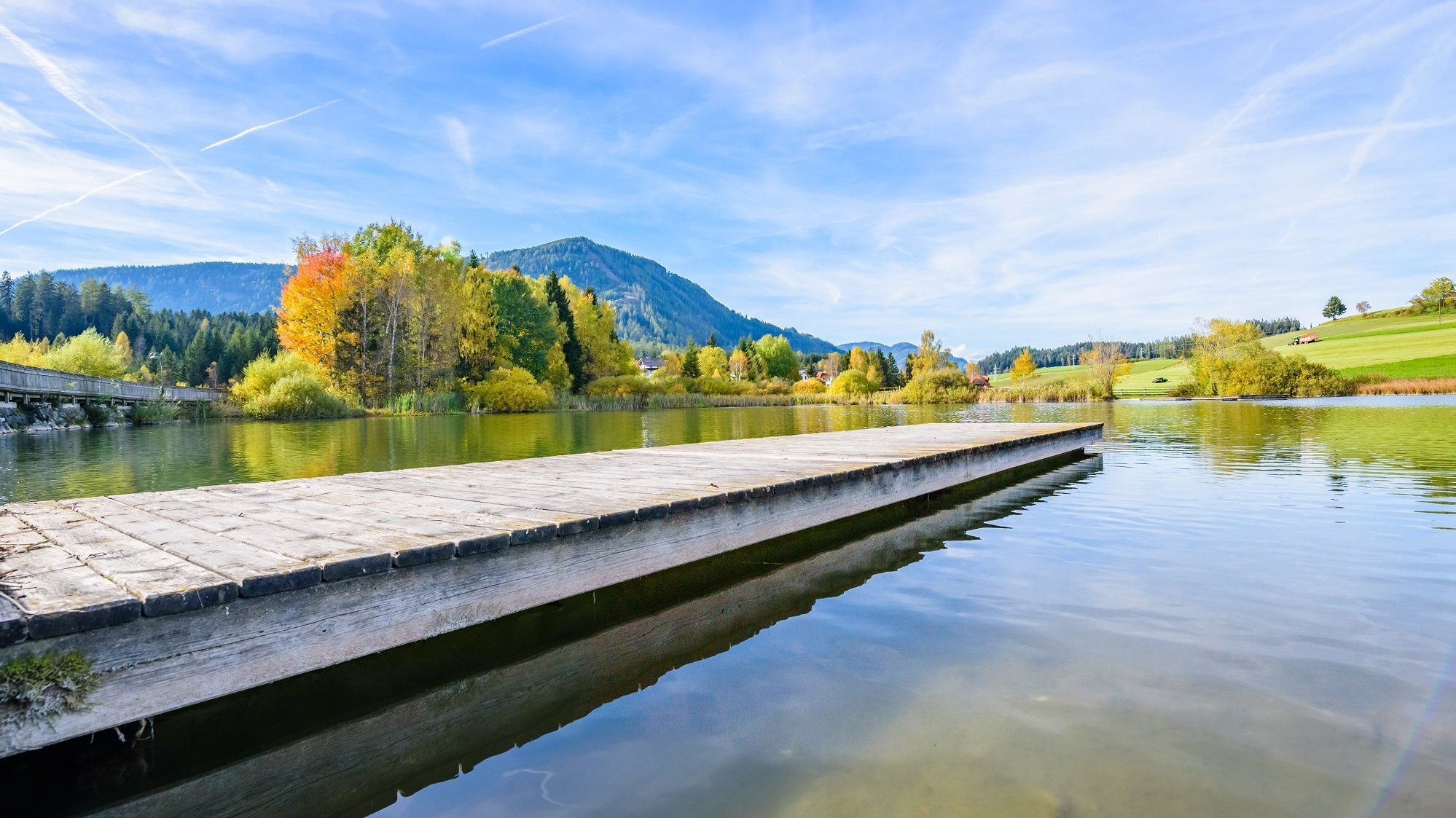 Herbstliche Idylle am See Ruhiger See umgeben von Bäumen mit Herbstlaub und einem Holzsteg unter blauem Himmel.