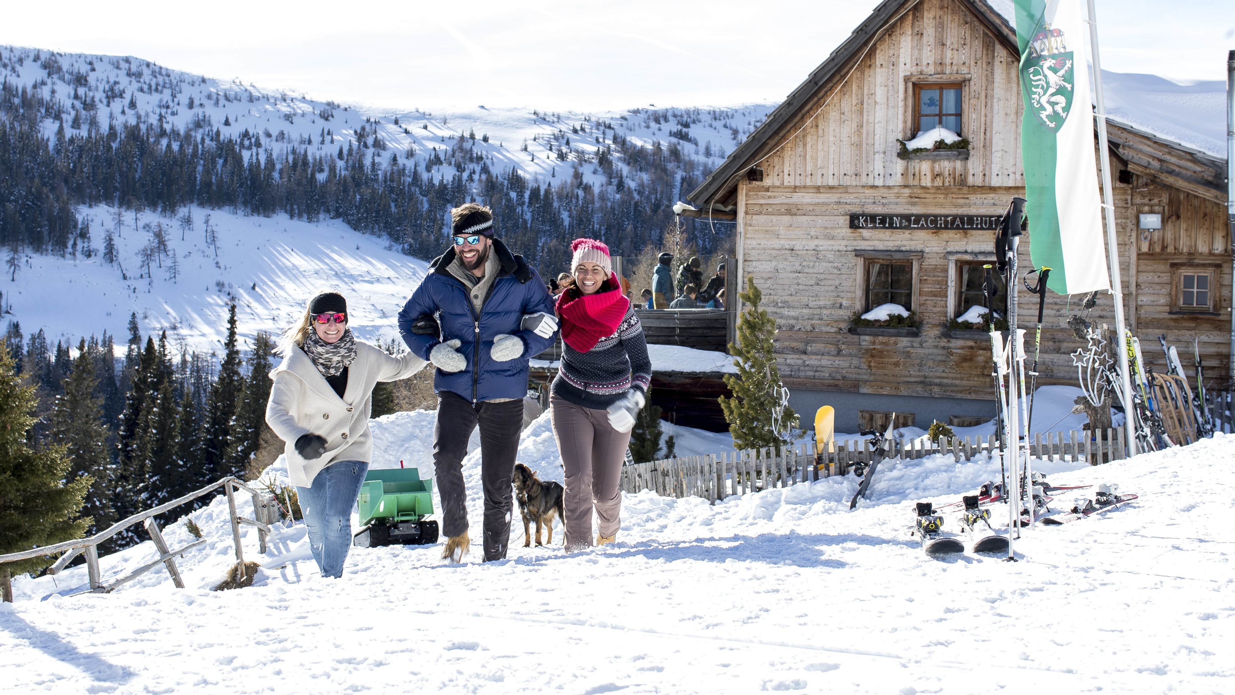 Drei Personen gehen durch eine verschneite Landschaft in der Steiermark zu einer Holzberghütte, Skiutensilien im Vordergrund.