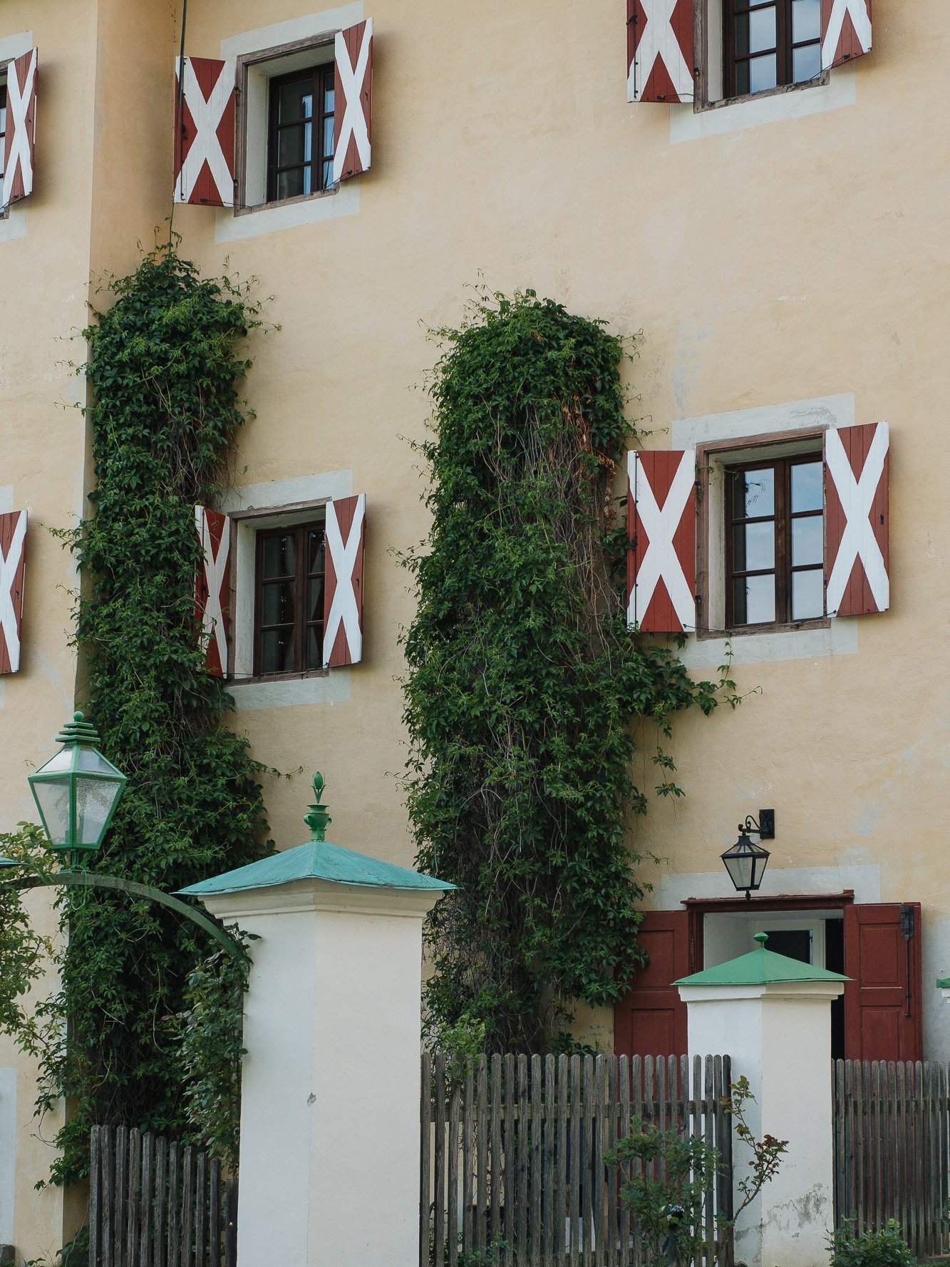 Fassade des Pichlschloss, mit roten und weißen Fensterläden, umgeben von Pflanzen und einem Holzzaun.