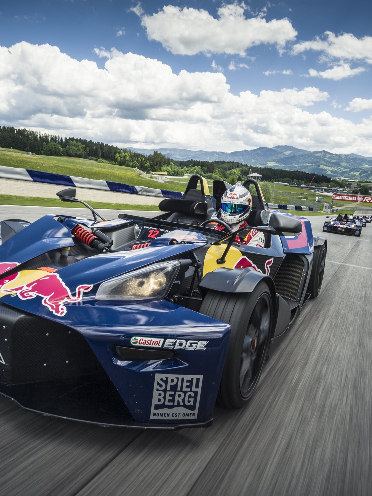 Rasante Fahrt auf der Rennstrecke des Red Bull Rings Ein Rennauto auf der Rennstrecke im Spielberg, mit Berglandschaft im Hintergrund und Wolken am Himmel.