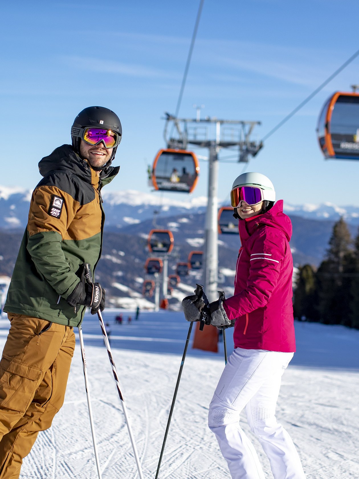 Zwei Skifahrer auf einer verschneiten Piste mit Skiliften und Bergen im Hintergrund.