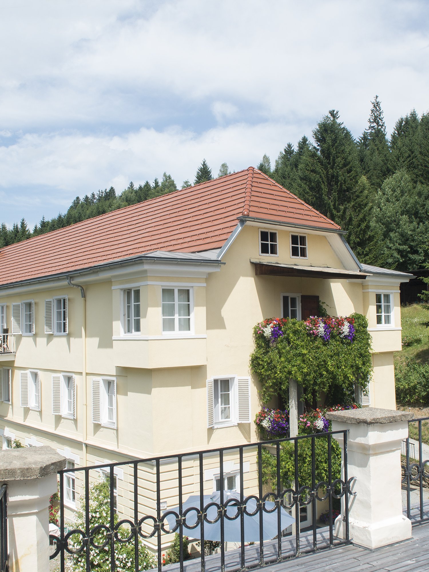 Mehrstöckiges Landhaus mit rotem Dach und blühenden Balkonen in einem waldreichen Umfeld