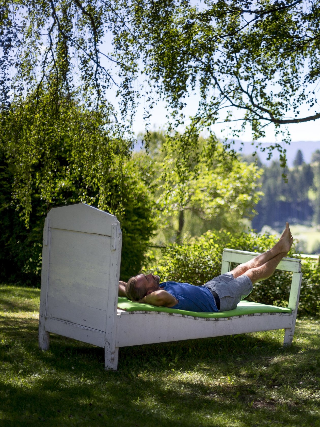 Person liegt entspannt auf einem Bett im Freien umgeben von Bäumen und Grünflächen.