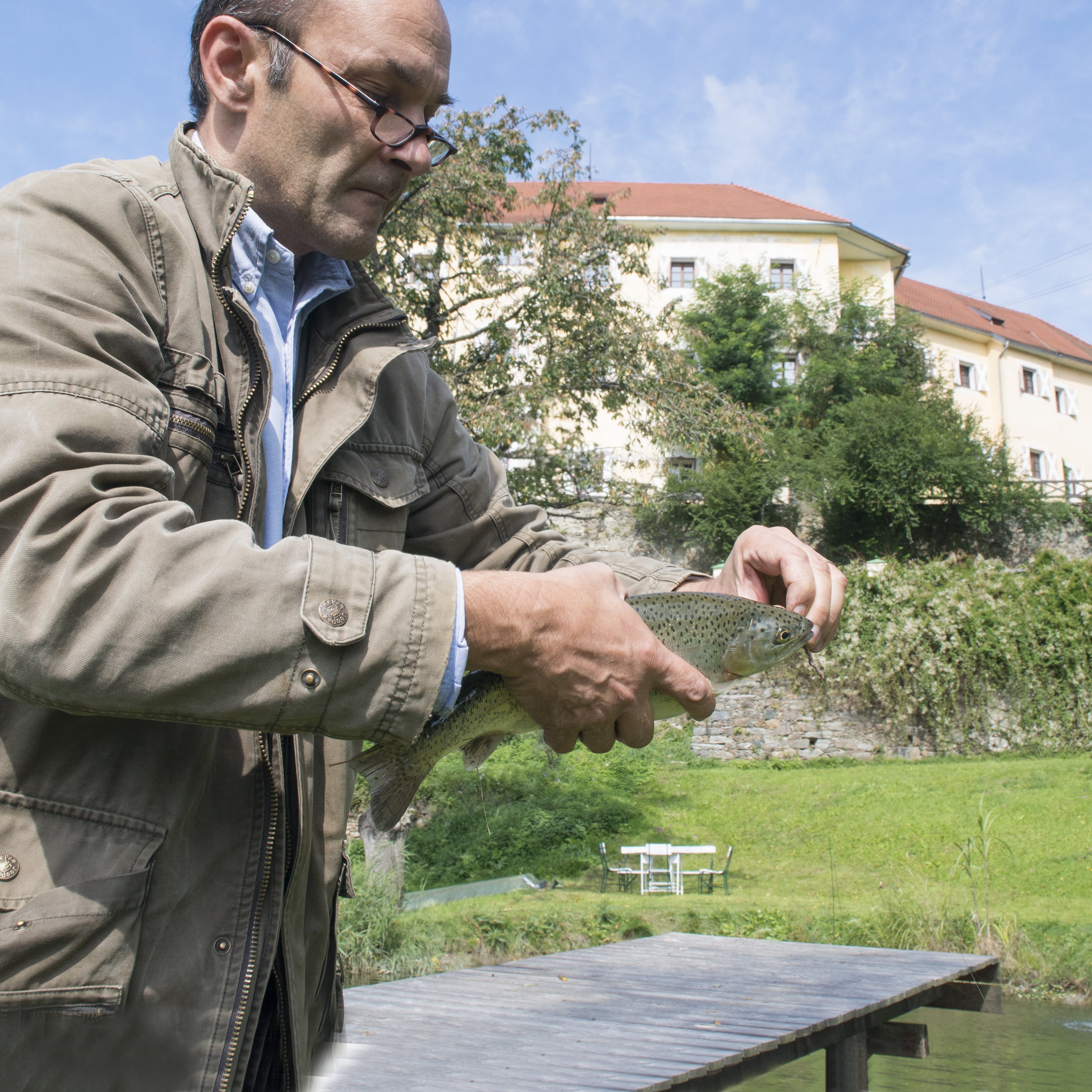 Fangfrische Forelle am Wasser Mann hält eine gefangene Forelle vor einem Gewässer mit einer grünen Landschaft im Hintergrund.