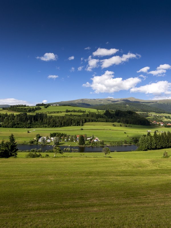 Landschaft mit Hügeln, einem klaren See und Bergen unter blauem Himmel mit Wolken