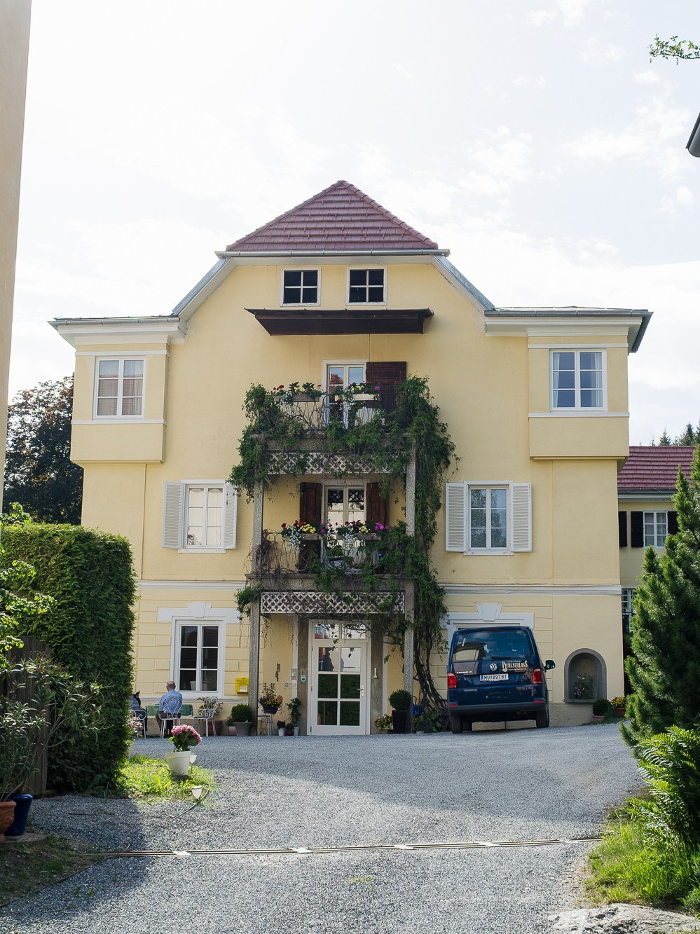 Zufahrt des Hotel Landsitz Pichlschloss Gelbes Wohngebäude mit Balkon und Pflanzen, umgeben von Garten und einer Kiesauffahrt.