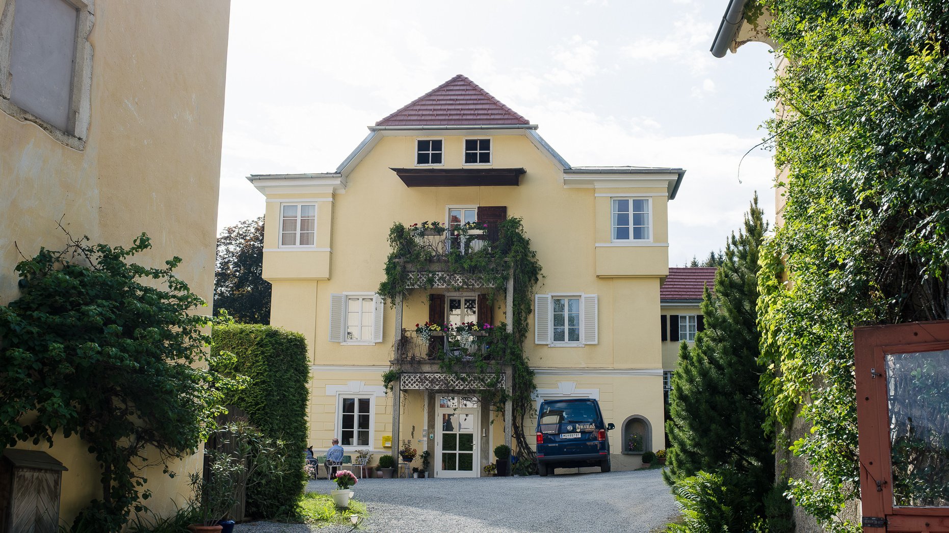 Charming Yellow House Surrounded by Nature Residential yellow building with balcony and green garden at the end of a gravel driveway