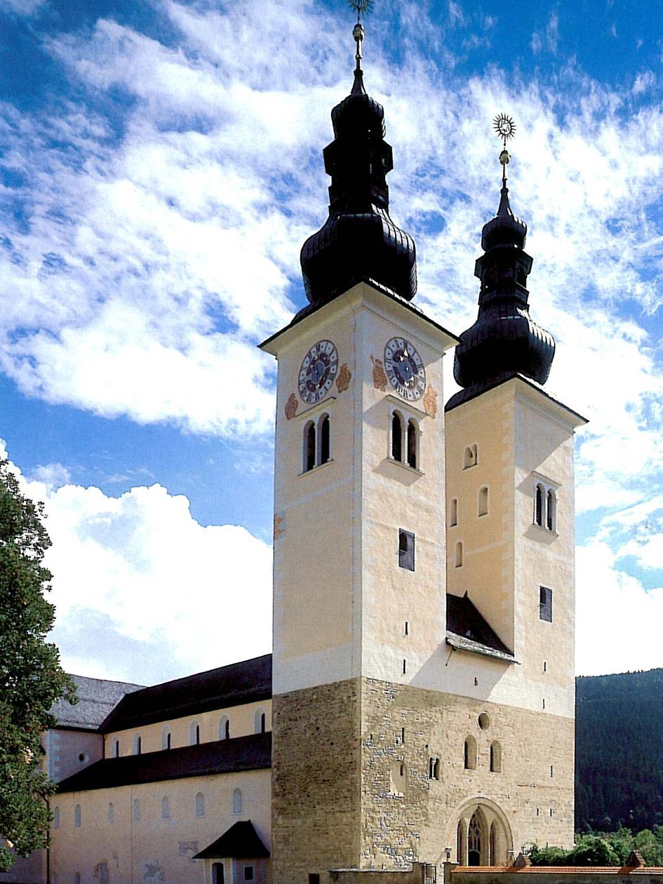 Historische Kirche mit beeindruckenden Türmen Historisches Gebäude einer Kirche mit zwei Türmen und Uhren, umgeben von Natur und Wolken.