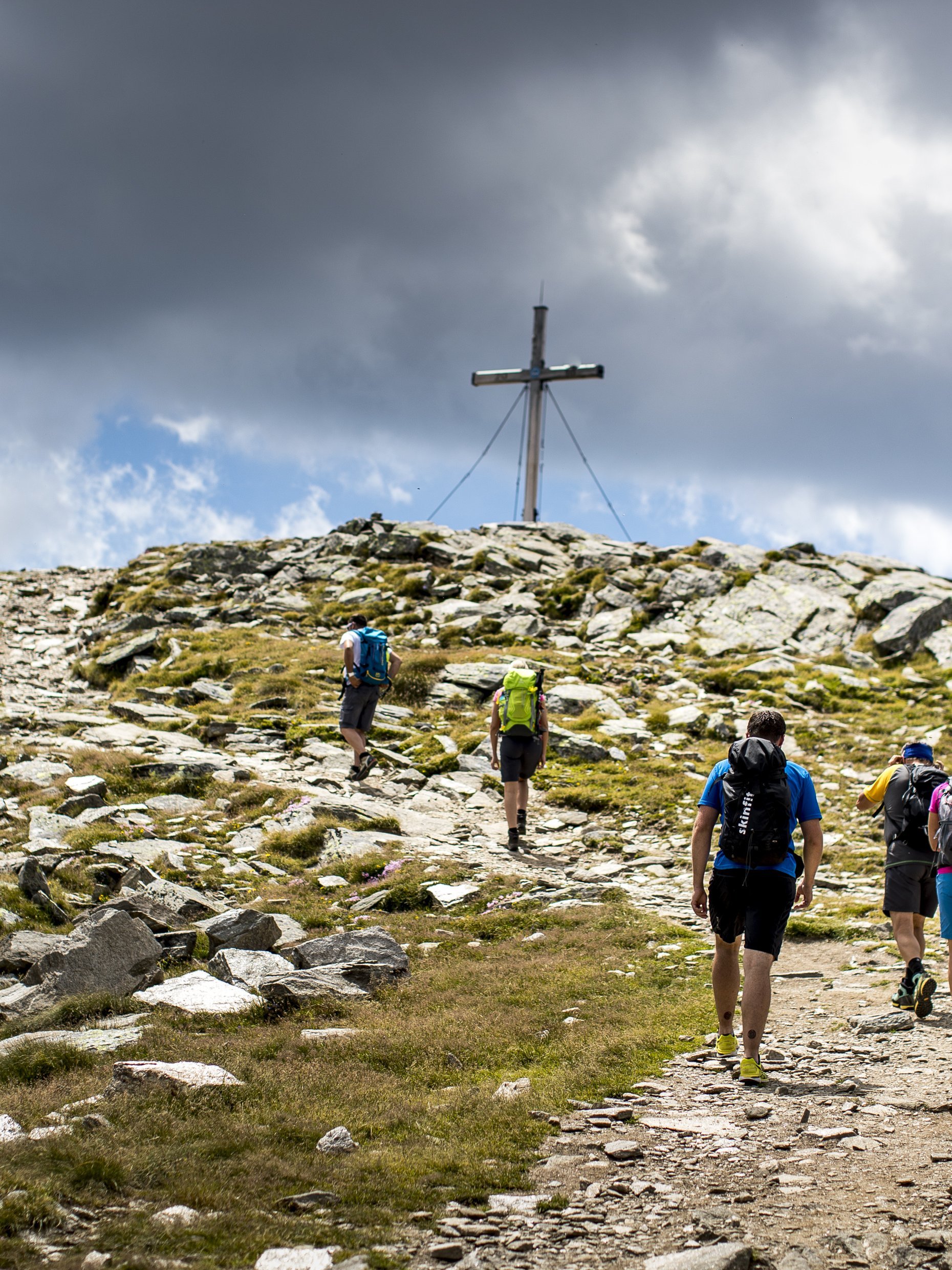 Hiking Adventure auf dem Zirbitzkogel Gruppe von Wanderern erklimmt einen steinigen Pfad zu einem Gipfel mit einem Kreuz.