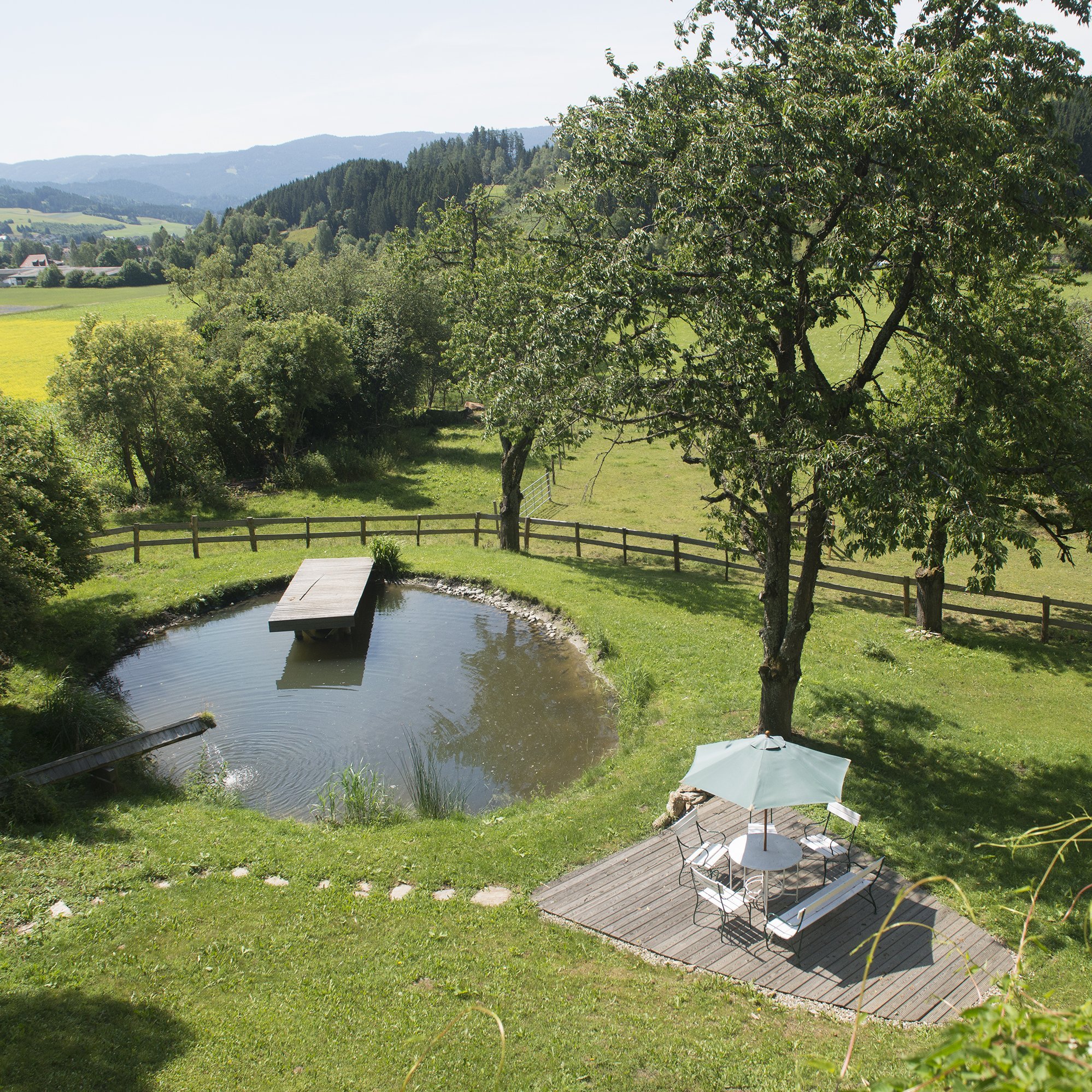 Idyllischer Teich mit Sitzbereich Runder Teich umgeben von Grün mit Holzplattform und Außensitzgelegenheiten unter einem Sonnenschirm.