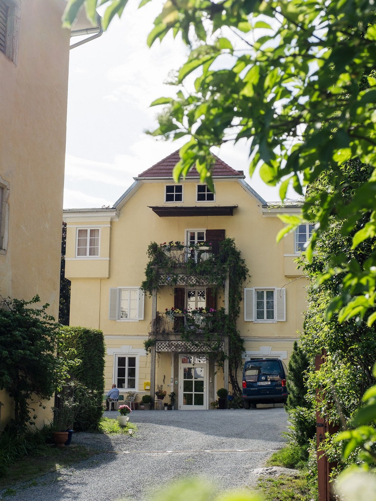 Zufahrt des Hotel Landsitz Pichlschloss Gelbes Wohngebäude mit Balkon und Pflanzen, umgeben von Grün und einem Kiesweg in einem Garten.