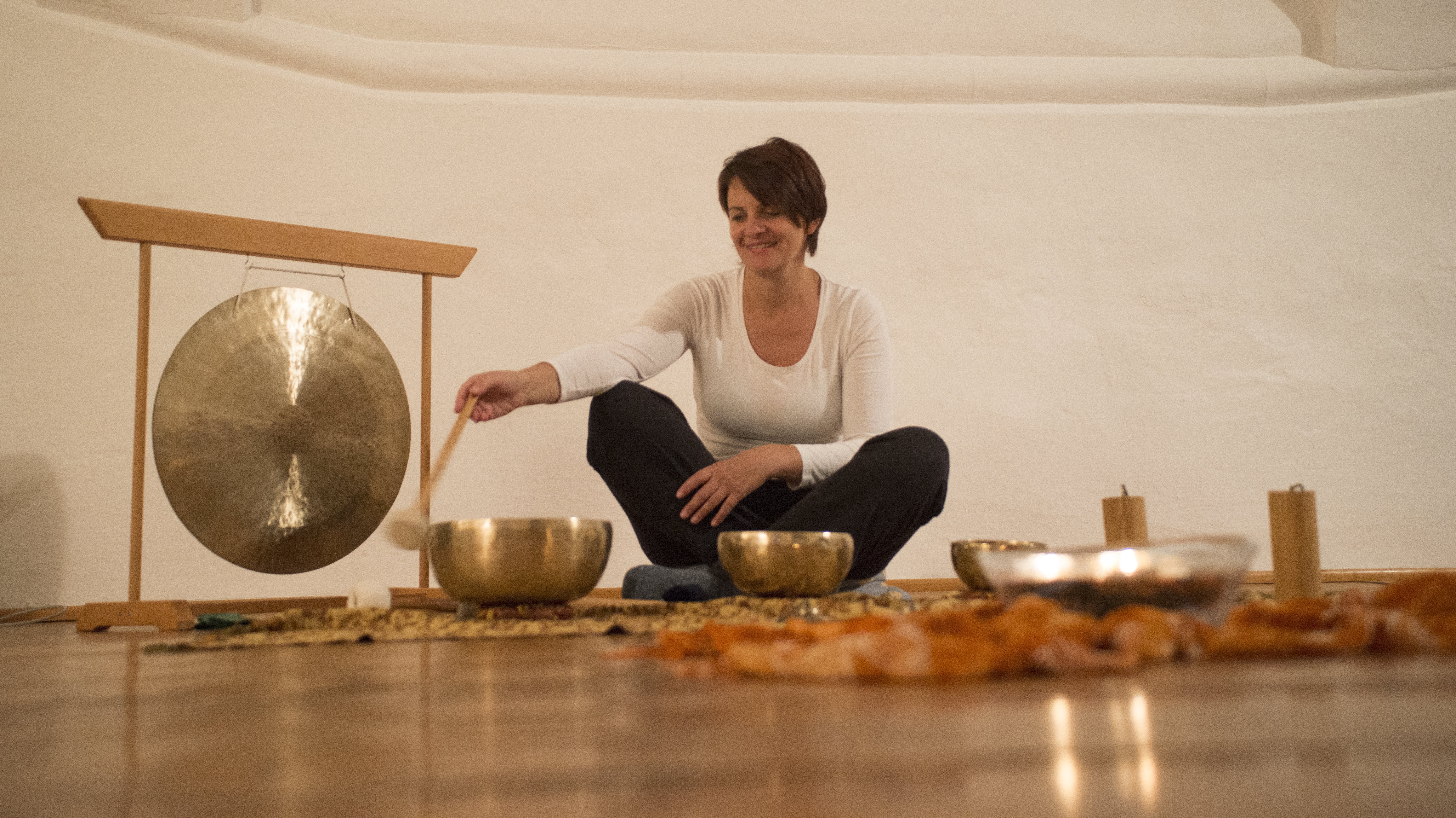Meditation mit Klangschalen Frau sitzt im Raum und spielt einen Gong, umgeben von Klangschalen und Kerzenlicht.