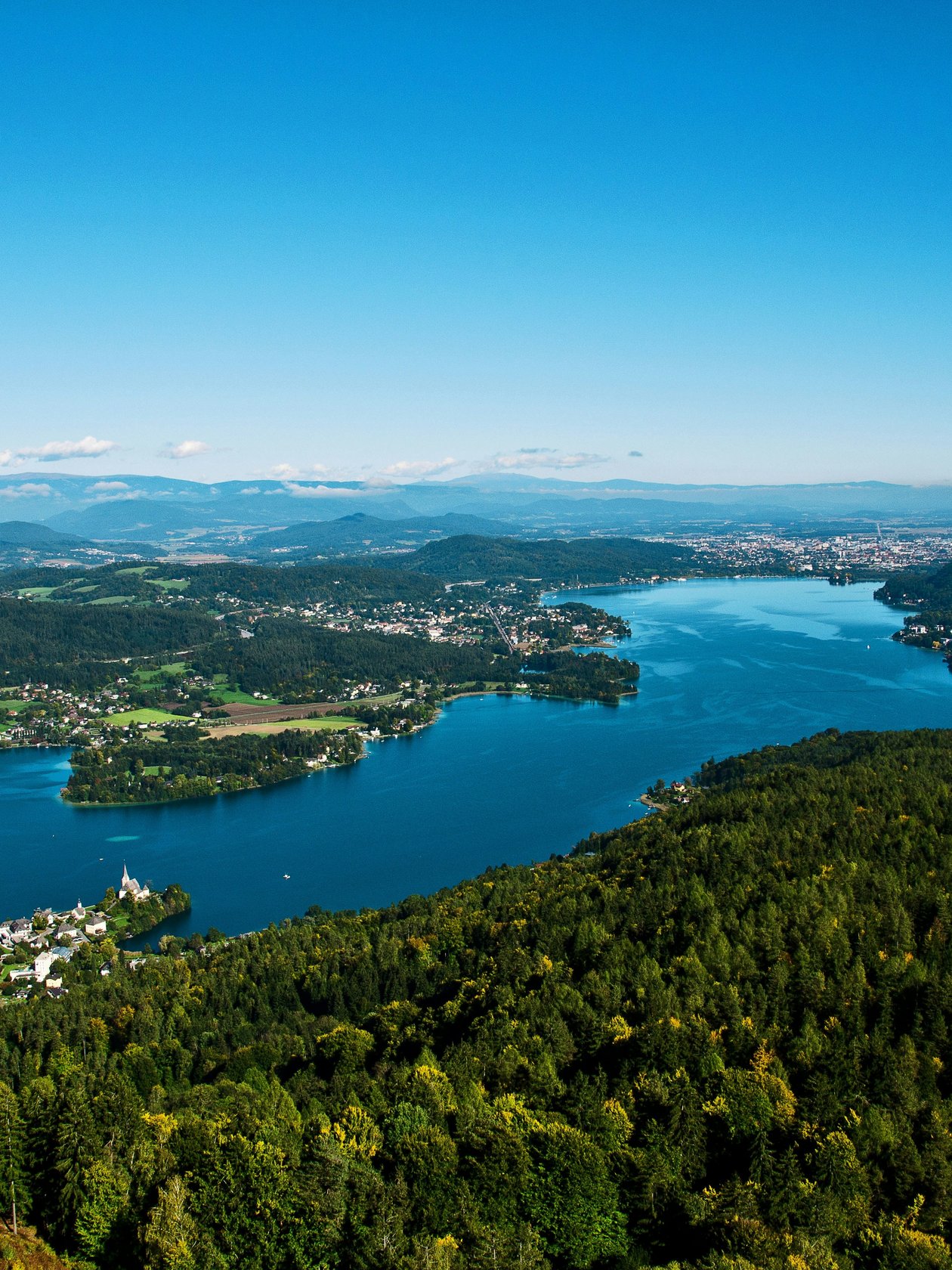 Panoramablick auf den Wörthersee Blick auf einen klaren See umgeben von Bergen und üppigem Grün unter blauem Himmel