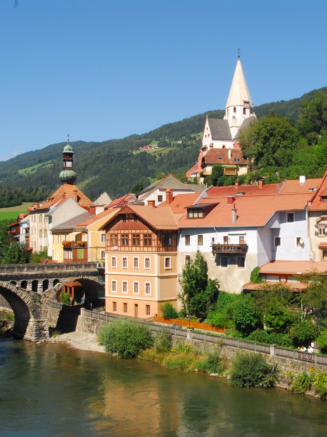 Malersches Dorf am Fluss Ein charmantes Dorf am Ufer eines Flusses mit historischen Gebäuden und Bergen im Hintergrund.