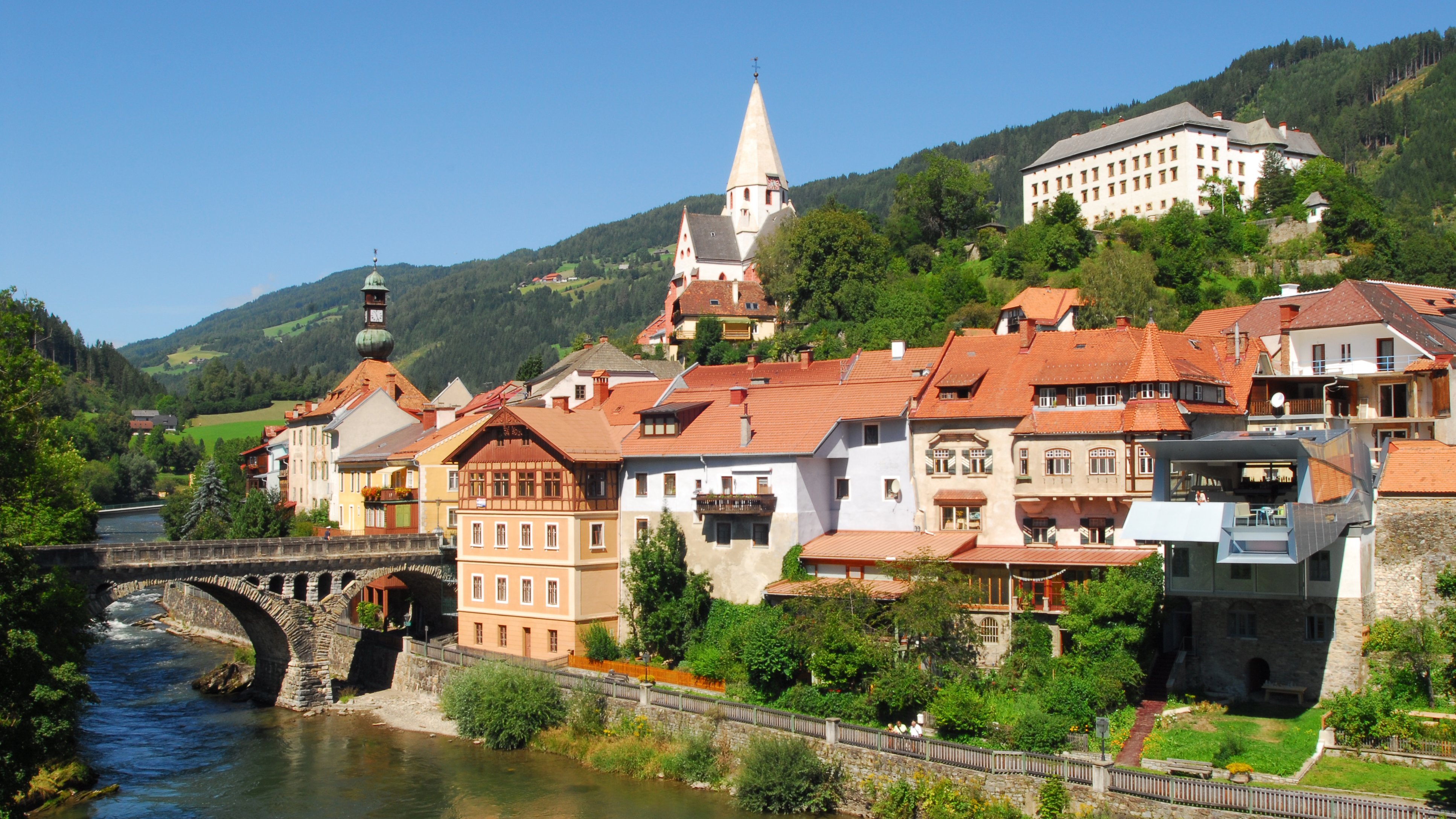 Malersches Dorf am Fluss Ein charmantes Dorf am Ufer eines Flusses mit historischen Gebäuden und Bergen im Hintergrund.