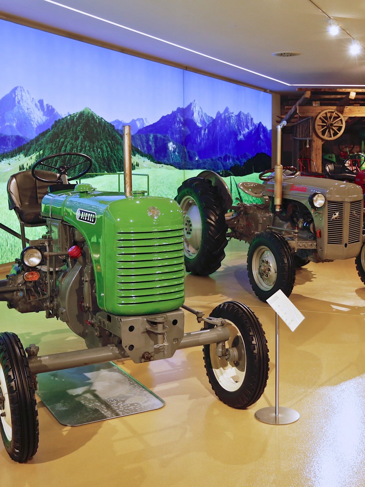 Drei historische Traktoren in einem Museum mit Berglandschaft auf großem Bildschirm im Hintergrund