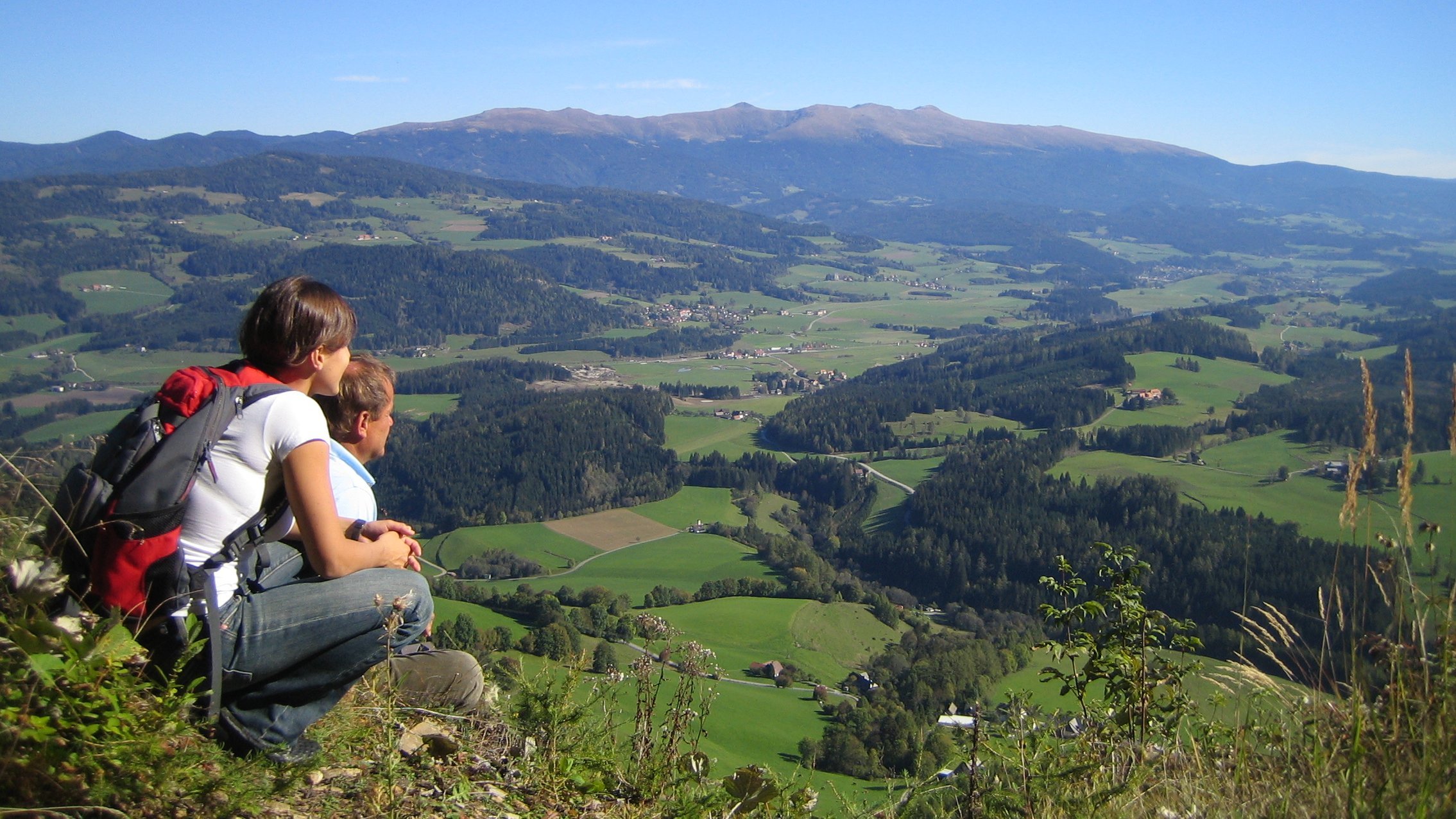 Breathtaking mountain view Two people sitting on a rocky ledge overlooking a green valley and mountains under a blue sky