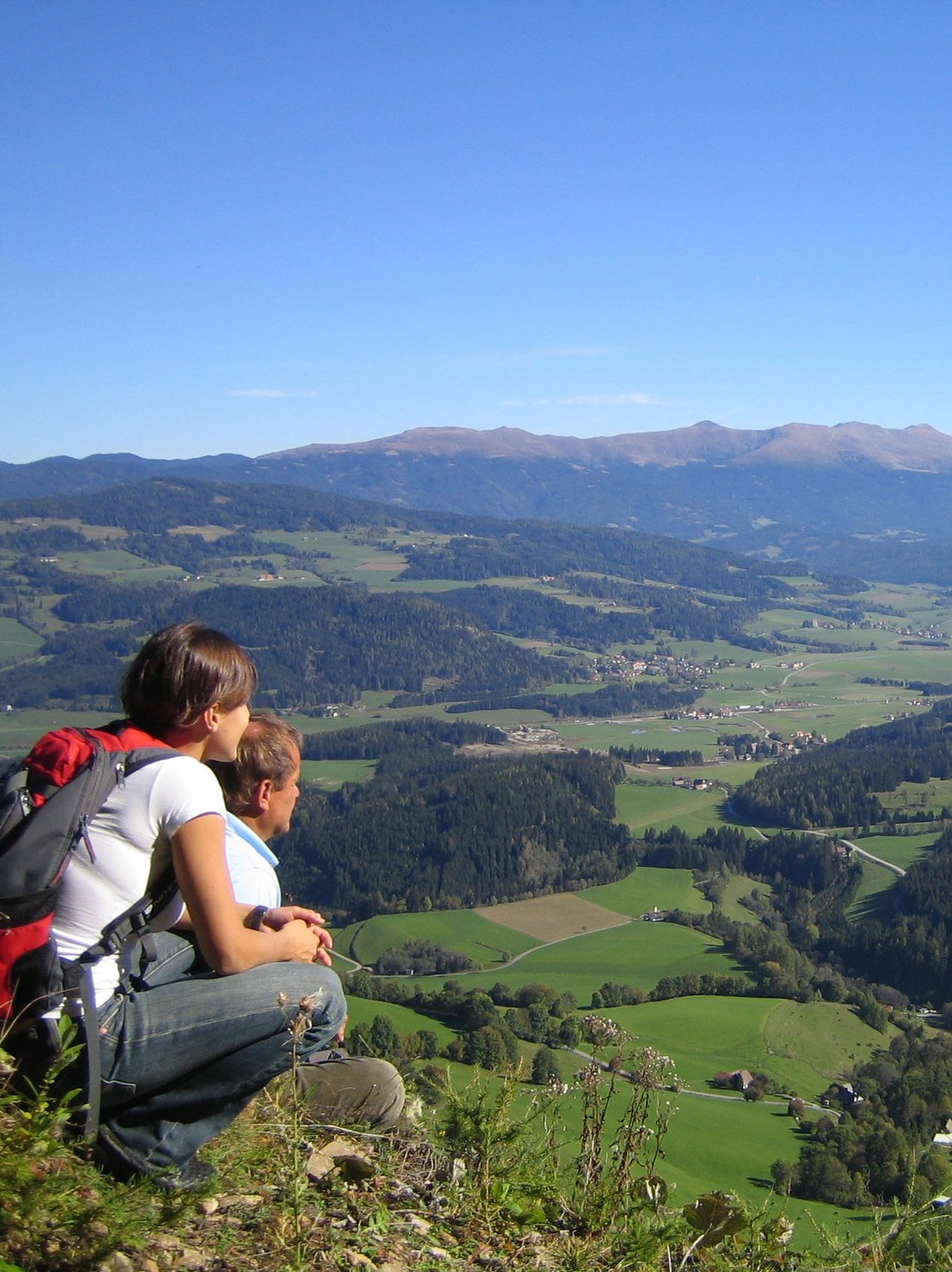Bergblick vom Felsen Zwei Personen sitzen auf einem felsigen Vorsprung und blicken auf eine grüne Landschaft mit Bergen im Hintergrund.