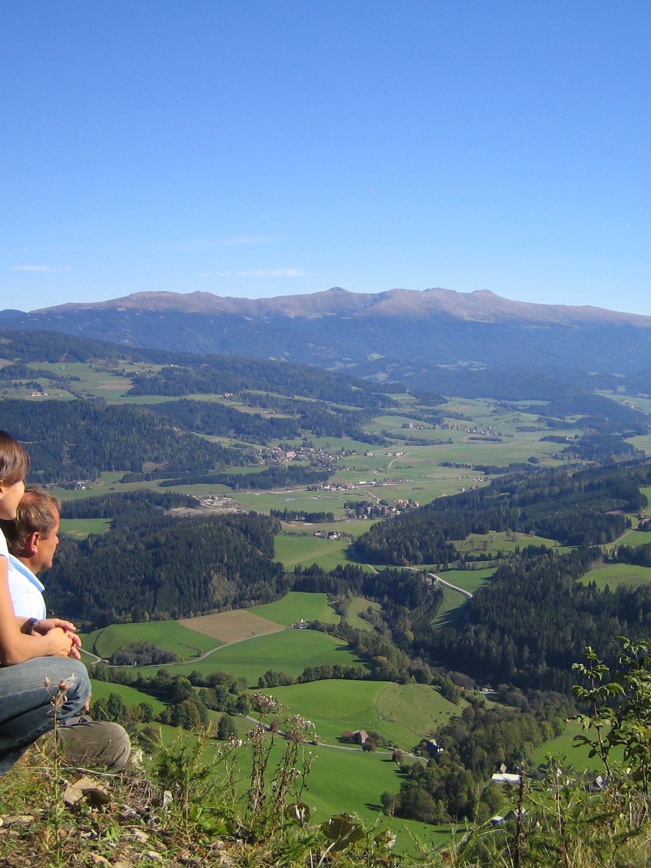 Bergblick vom Felsen Zwei Personen sitzen auf einem felsigen Vorsprung und blicken auf eine grüne Landschaft mit Bergen im Hintergrund.