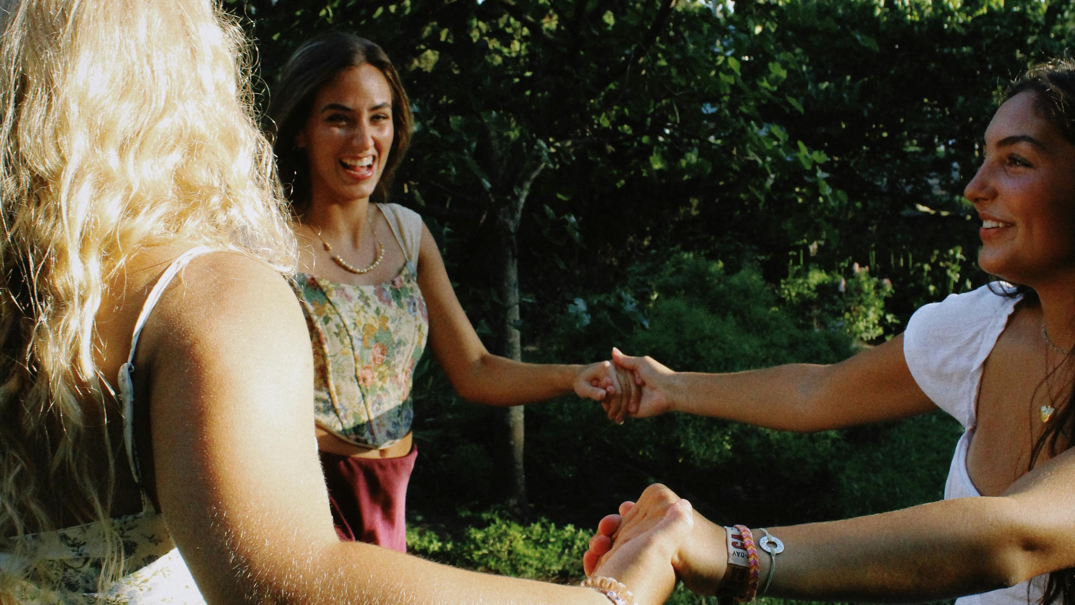 Freundschaft in der Natur Drei Frauen tanzen Hand in Hand in einer grünen Umgebung unter freiem Himmel. Sie strahlen Freude aus.