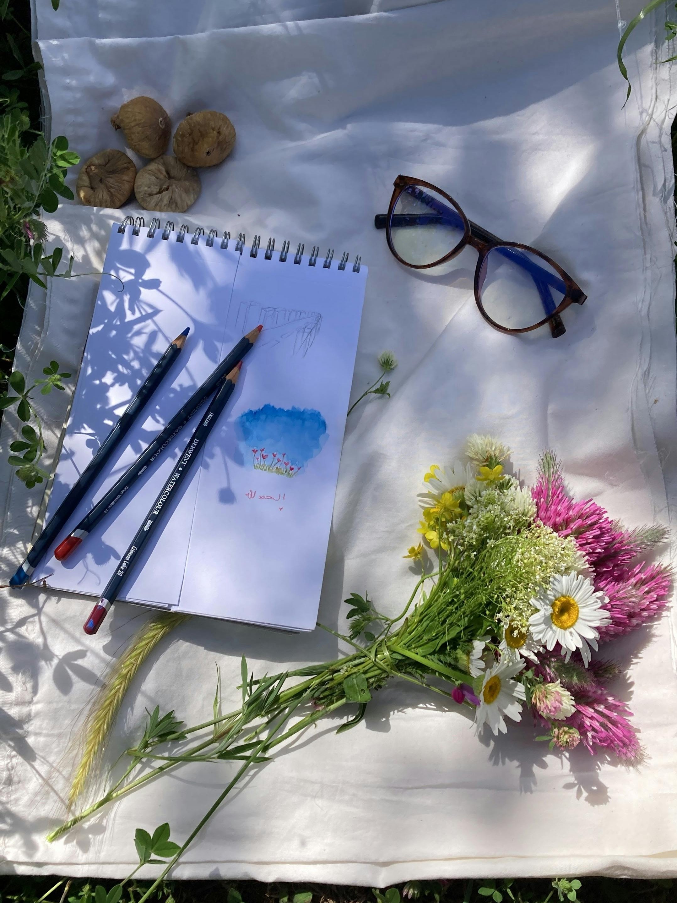 Kreativität im Freien Flat Lay mit Zeichenutensilien, Skizzenbuch, Blumenstrauß und Sonnenbrille auf weißem Stoff