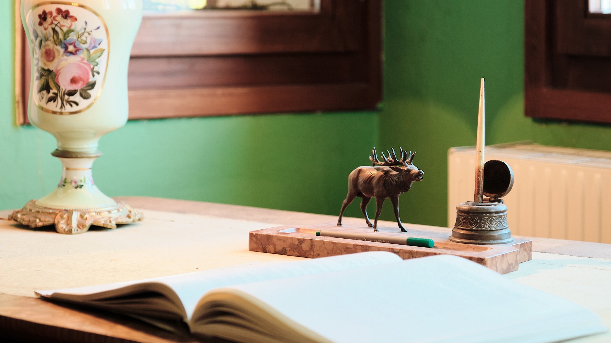 Vintage Schreibtischstil für kreative Köpfe Ein Schreibtisch mit dekorativer Lampe, Elchfigur, Feder und offenem Notizbuch vor grüner Wand