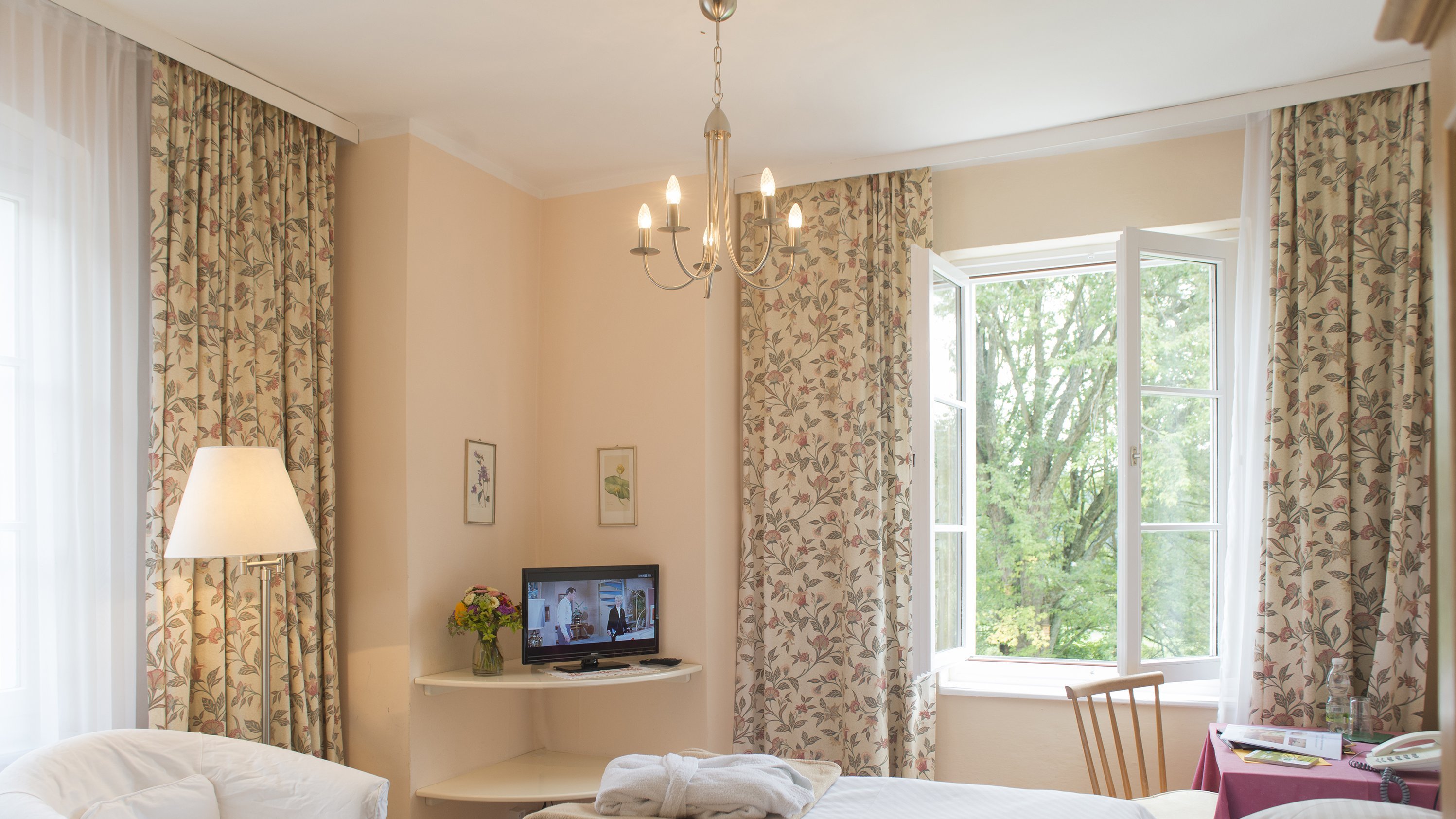 Hotelzimmer mit einem Bett, Stuhl und Fernseher sowie großen Fenstern und Blumenvorhängen