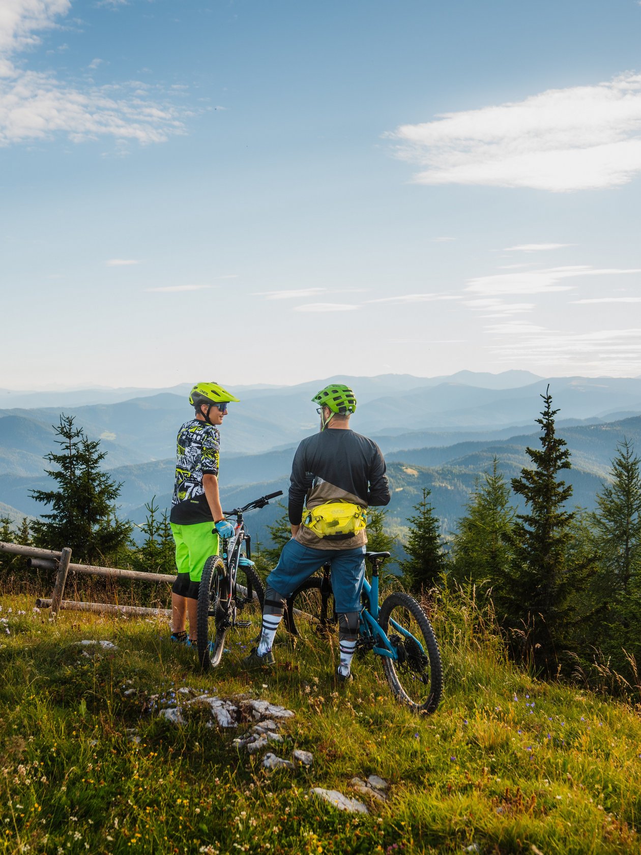 Abenteuer auf dem Grebenzen Trail Zwei Radfahrer stehen auf einem Hügel und blicken auf eine Berglandschaft mit Bäumen und klarem Himmel.