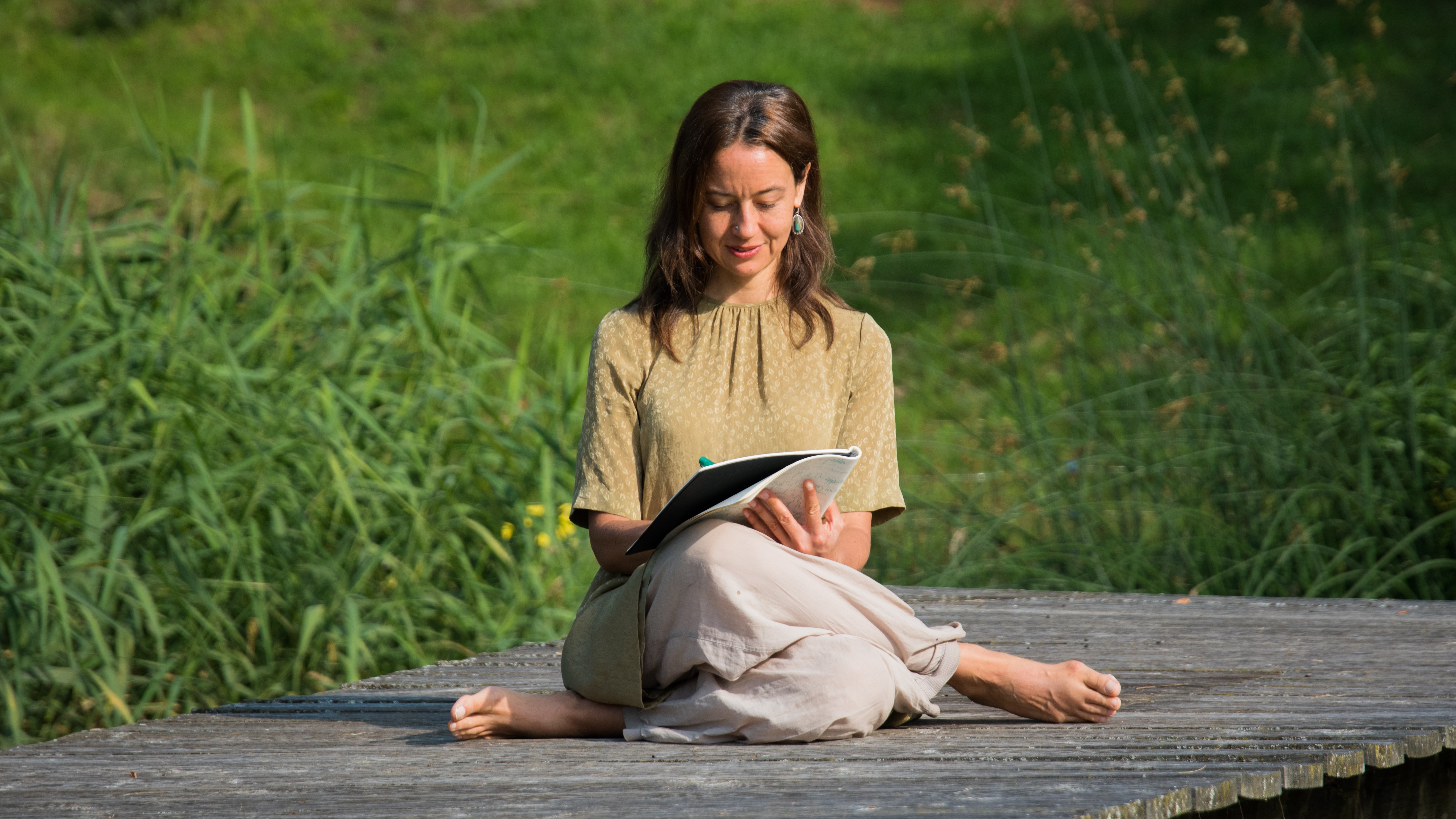 Schreibende Frau in der Natur Eine Frau sitzt im Freien auf einem Holzplatz und schreibt in ein Notizbuch, umgeben von Pflanzen.