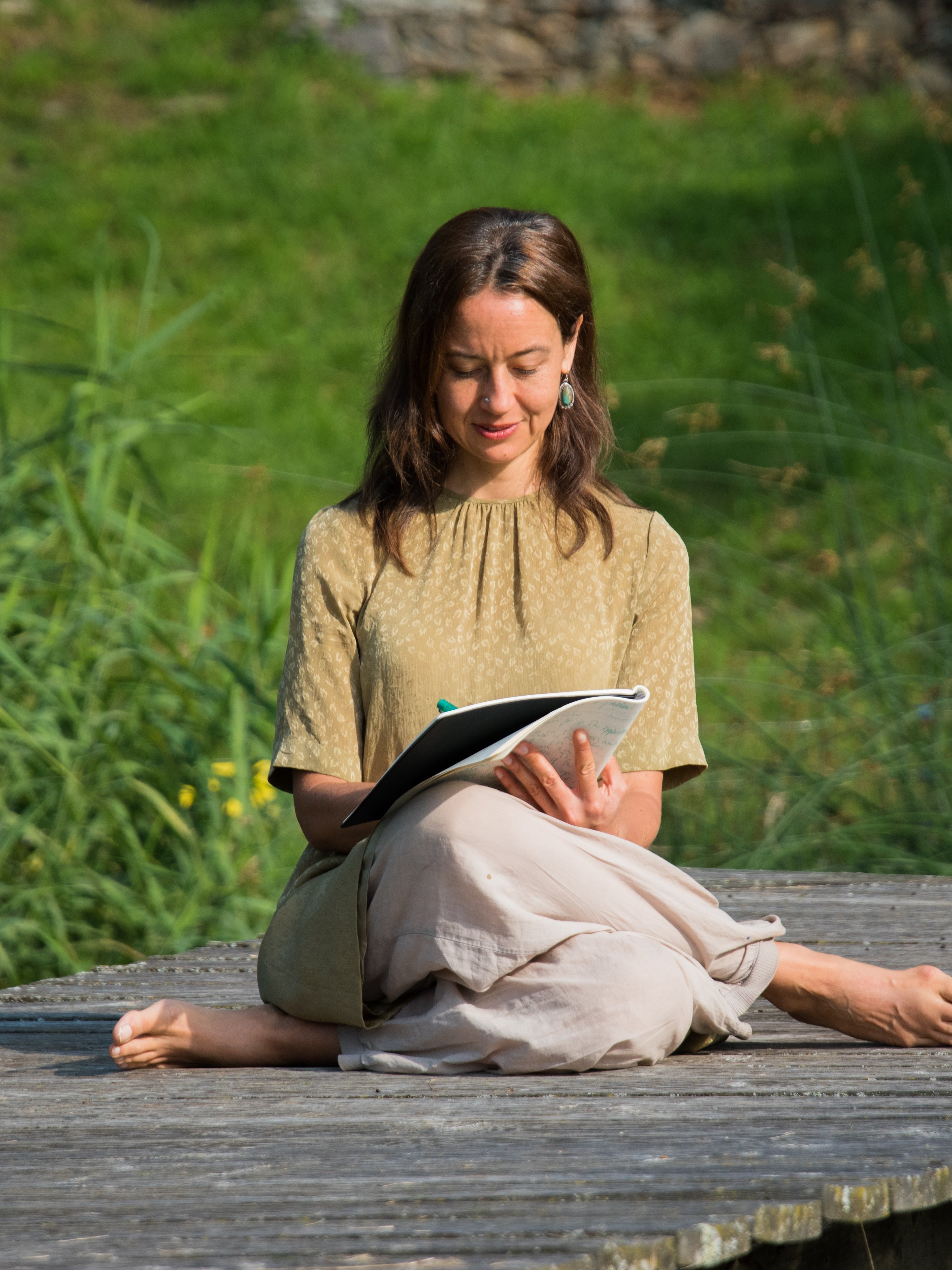 Mindful Writing in Nature A woman sits cross-legged on a wooden platform writing in a notebook surrounded by trees and greenery.
