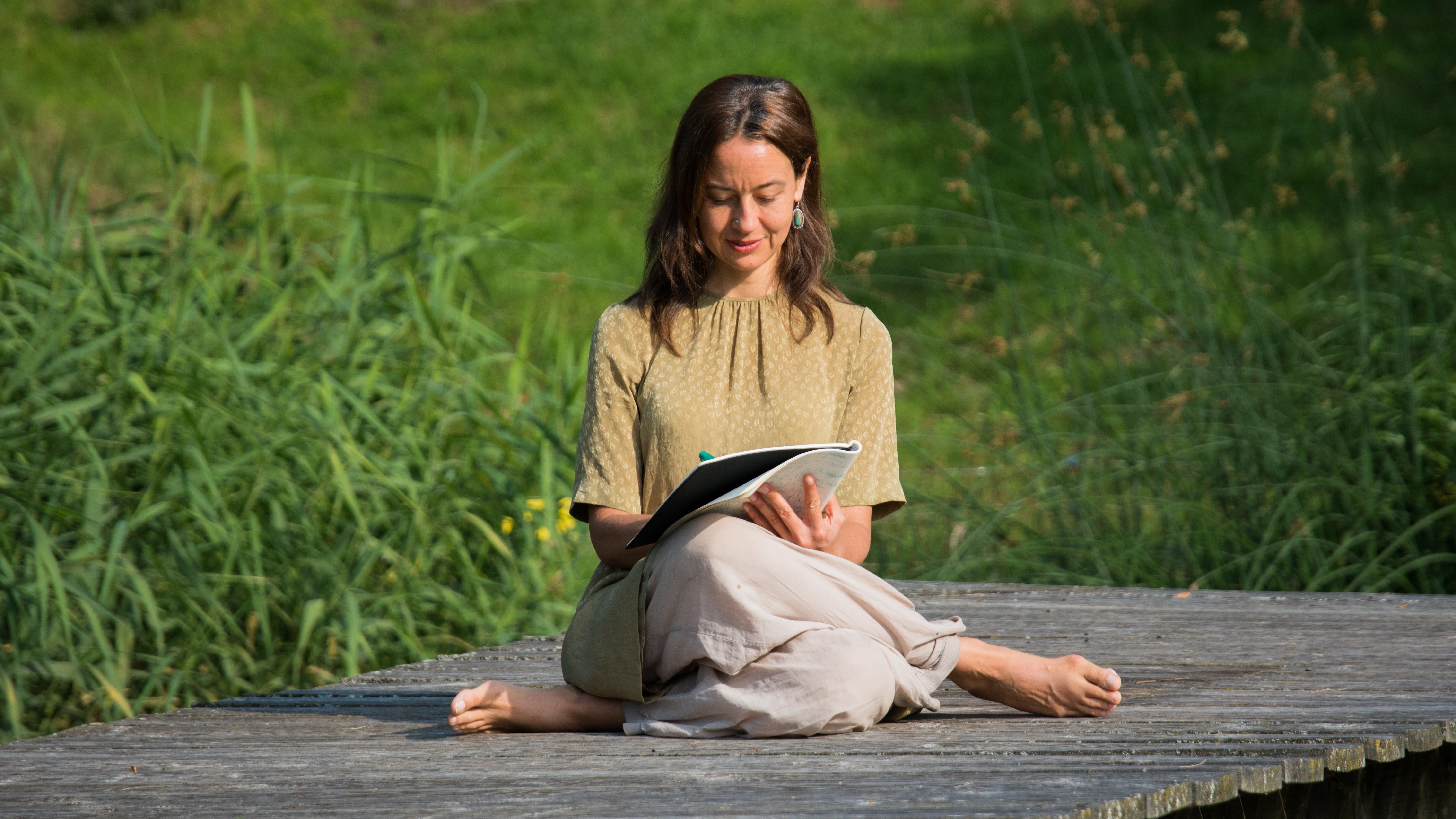 Schreibende Frau in der Natur Eine Frau sitzt im Freien auf einem Holzplatz und schreibt in ein Notizbuch, umgeben von Pflanzen.