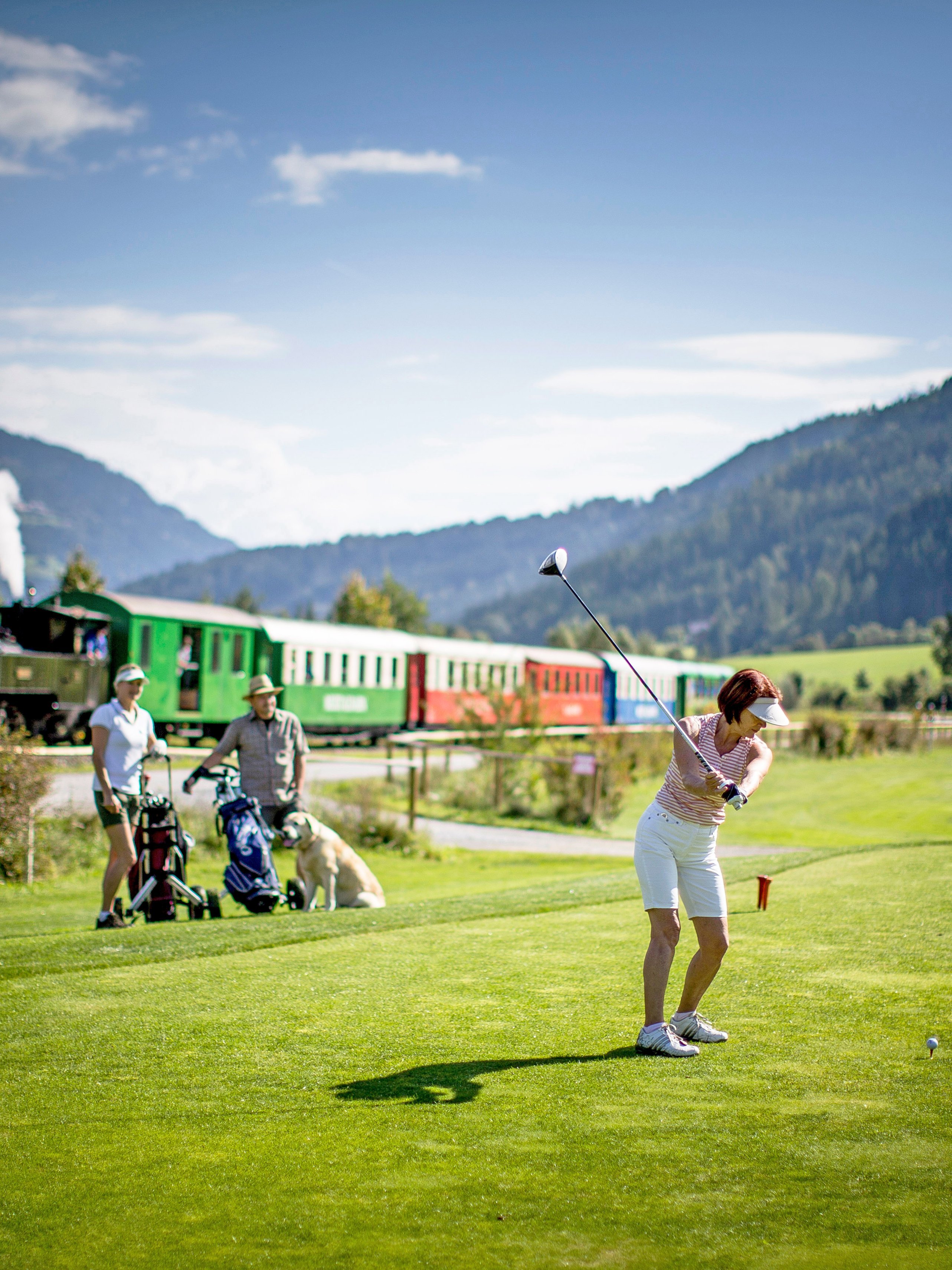 Golfspiel im Naturparadies Golfer auf einem Golfplatz mit einem bunten Zug im Hintergrund, umgeben von Bergen und Bäumen.