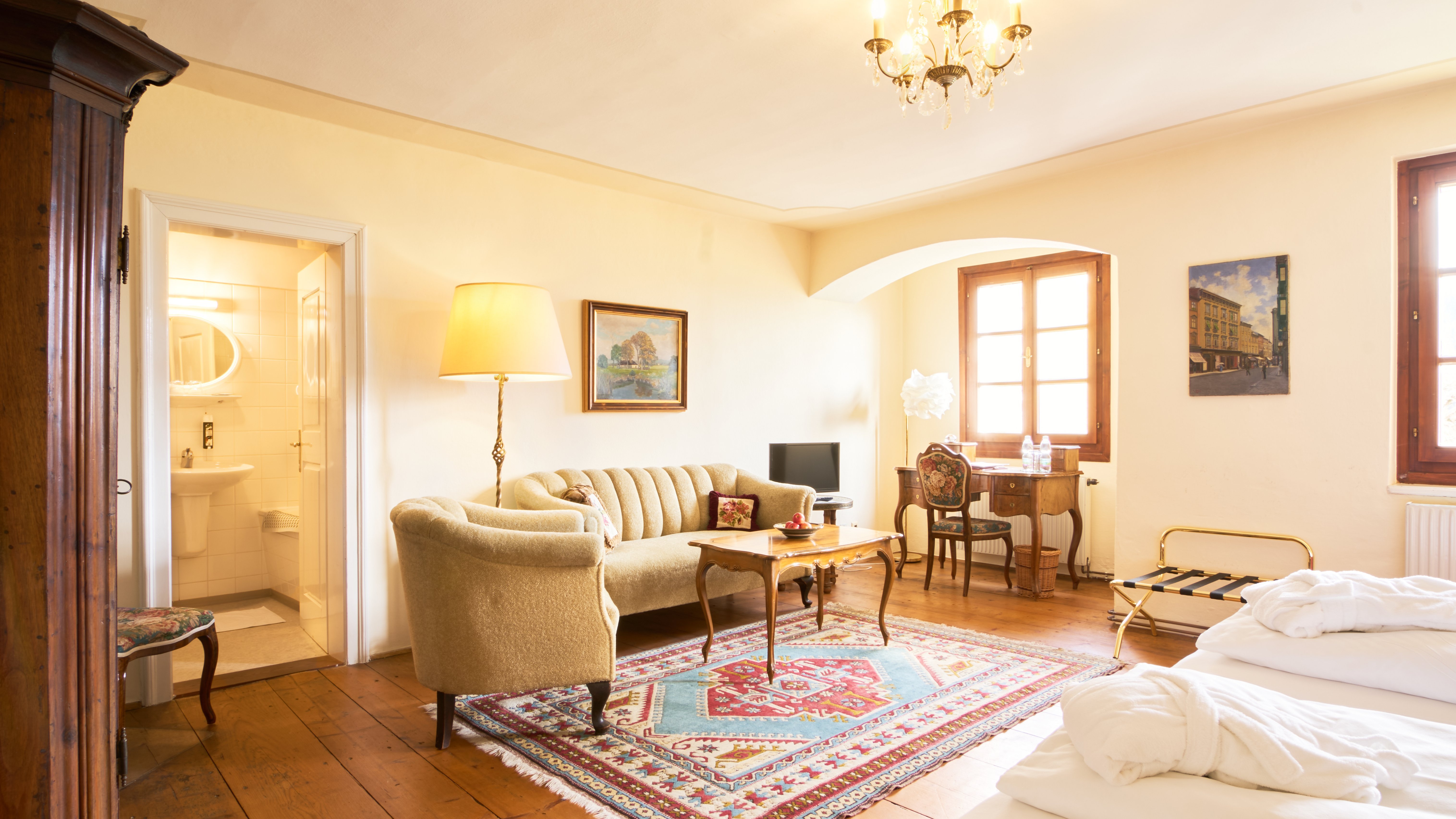 Elegante Zimmergestaltung im Hotel Landsitz Pischlschloss Behagliches Hotelzimmer mit Sofa, Tisch und Bett in weißer Bettwäsche sowie angrenzendem Badezimmer.